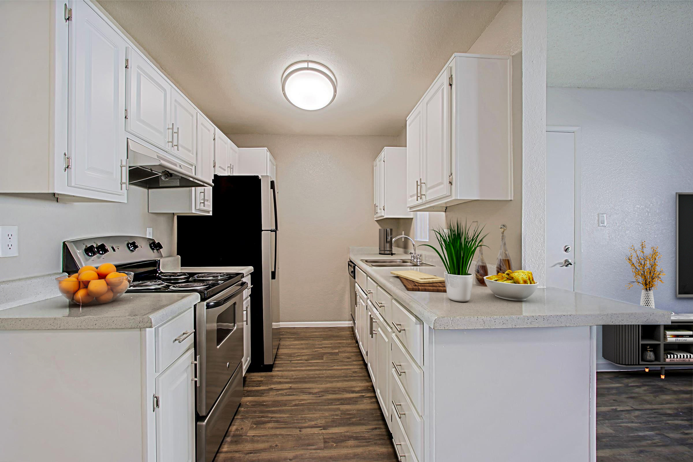 A modern kitchen featuring white cabinets, a stainless steel stove, and refrigerator. Countertops are light gray with a bowl of oranges, a small plant, and a dish of snacks. The space is well-lit with a ceiling light, and the floor has a wood-like appearance, creating a warm and inviting atmosphere.
