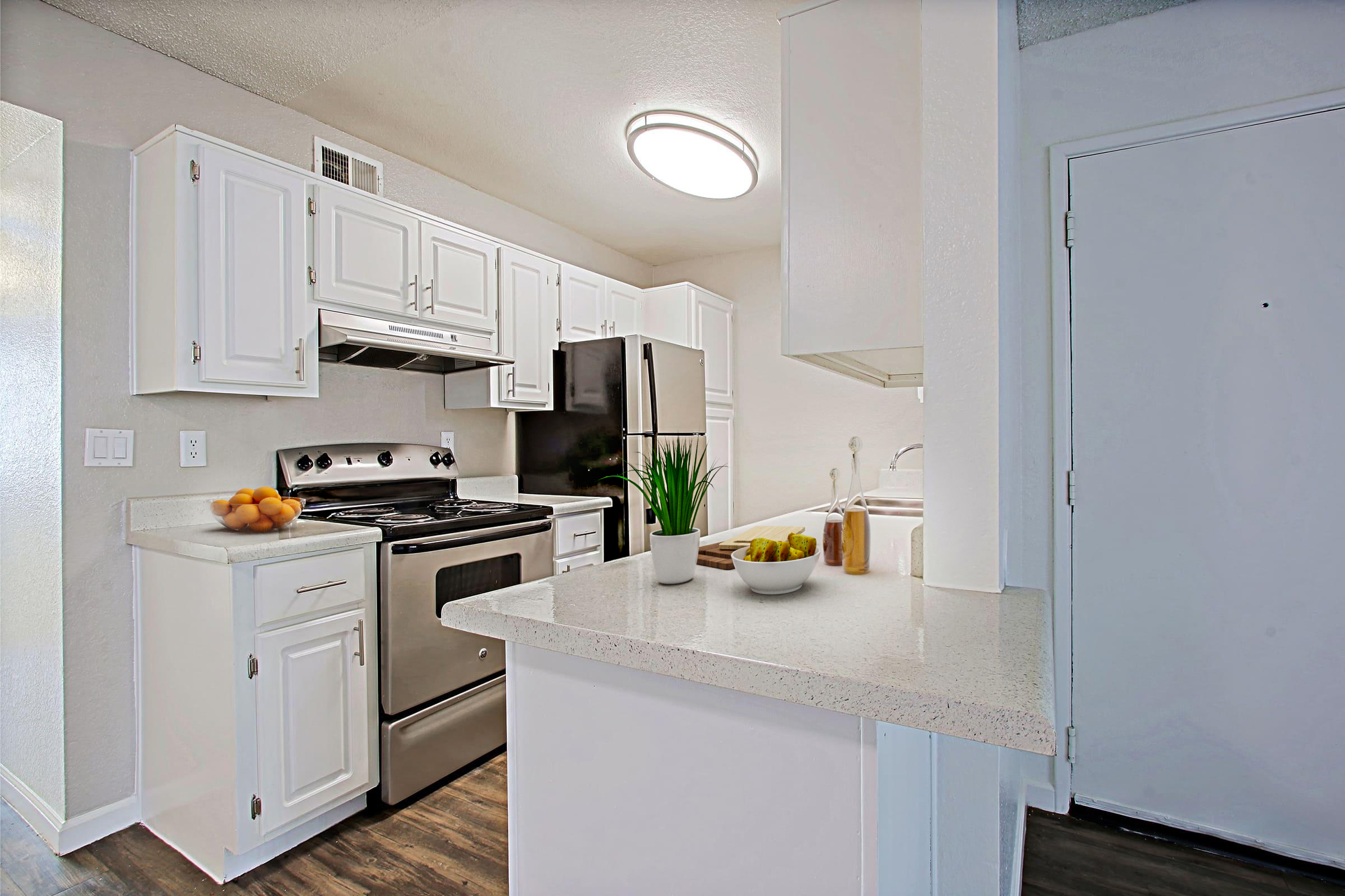 Modern kitchen featuring white cabinets, a black refrigerator, and stainless steel appliances. A countertop with a bowl of fruit and a potted plant is visible, along with a washer and dryer. Bright lighting enhances the spacious, clean atmosphere.