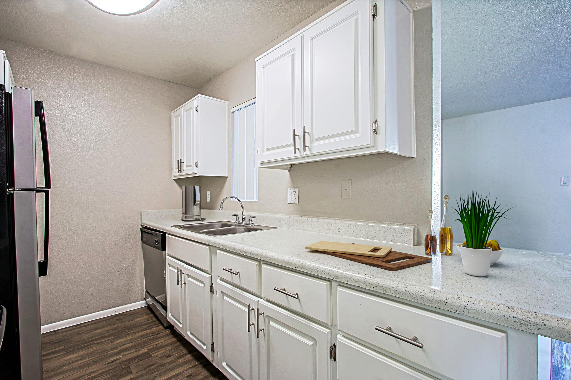 Modern kitchen featuring white cabinets, a stainless steel refrigerator, and a sleek countertop. The space includes a sink, dishwasher, and a wooden cutting board, with a small plant accentuating the decor. Natural light fills the room through a nearby window, enhancing the bright and clean atmosphere.