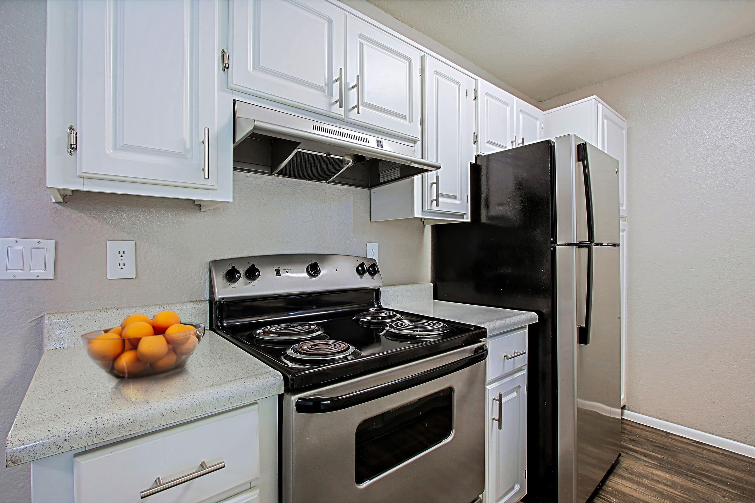 A modern kitchen featuring white cabinets, a black stove with four burners, a stainless steel refrigerator, and a countertop with a bowl of oranges. The walls are painted in a light color, and the flooring appears to be dark wood.