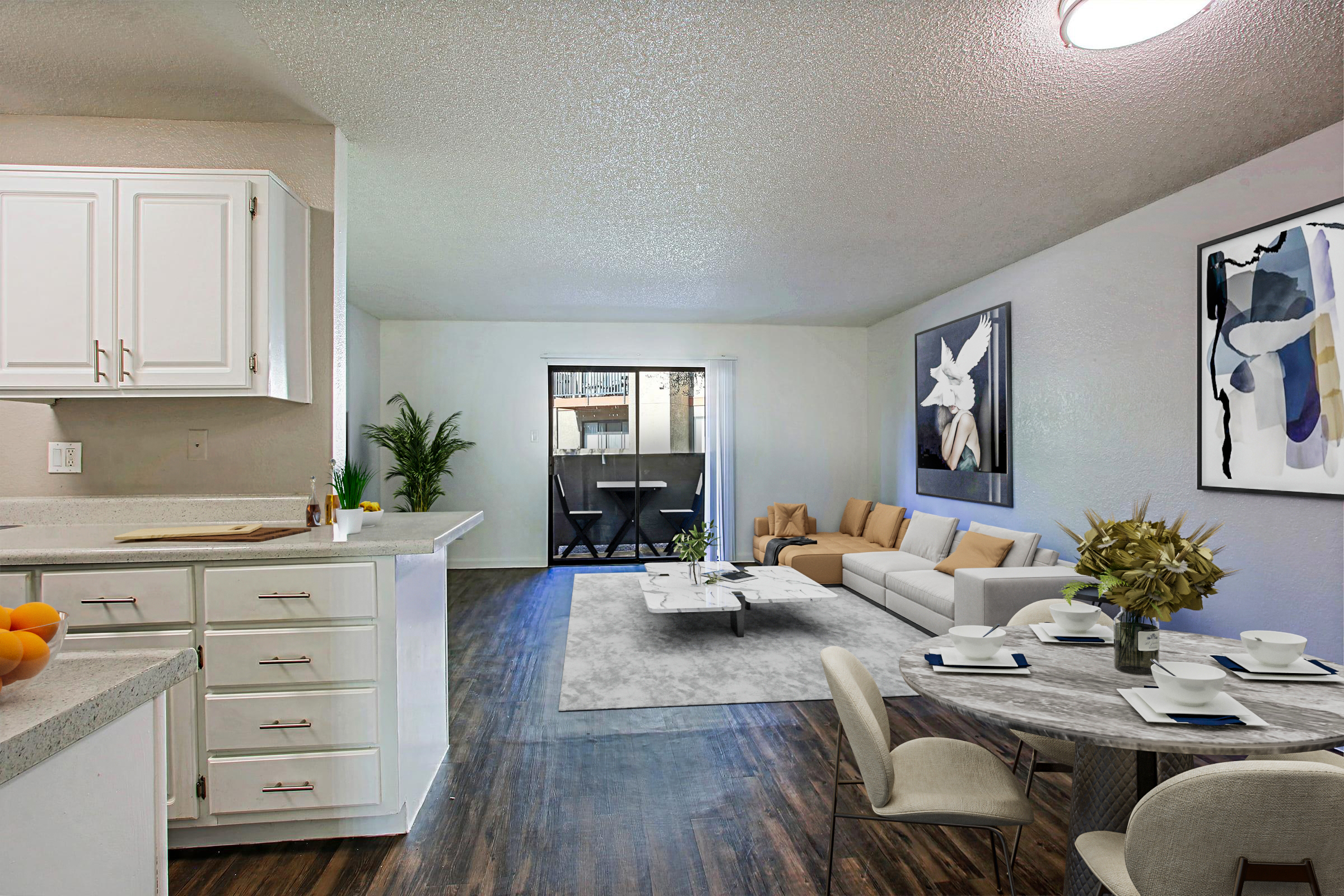 A modern living space featuring a light-filled open layout. The kitchen with white cabinetry is adjacent to a cozy living area with a beige sofa and a coffee table. A dining area with a round table is set for two. Large windows offer a view of a small balcony. Decorative plants and art enhance the aesthetic.