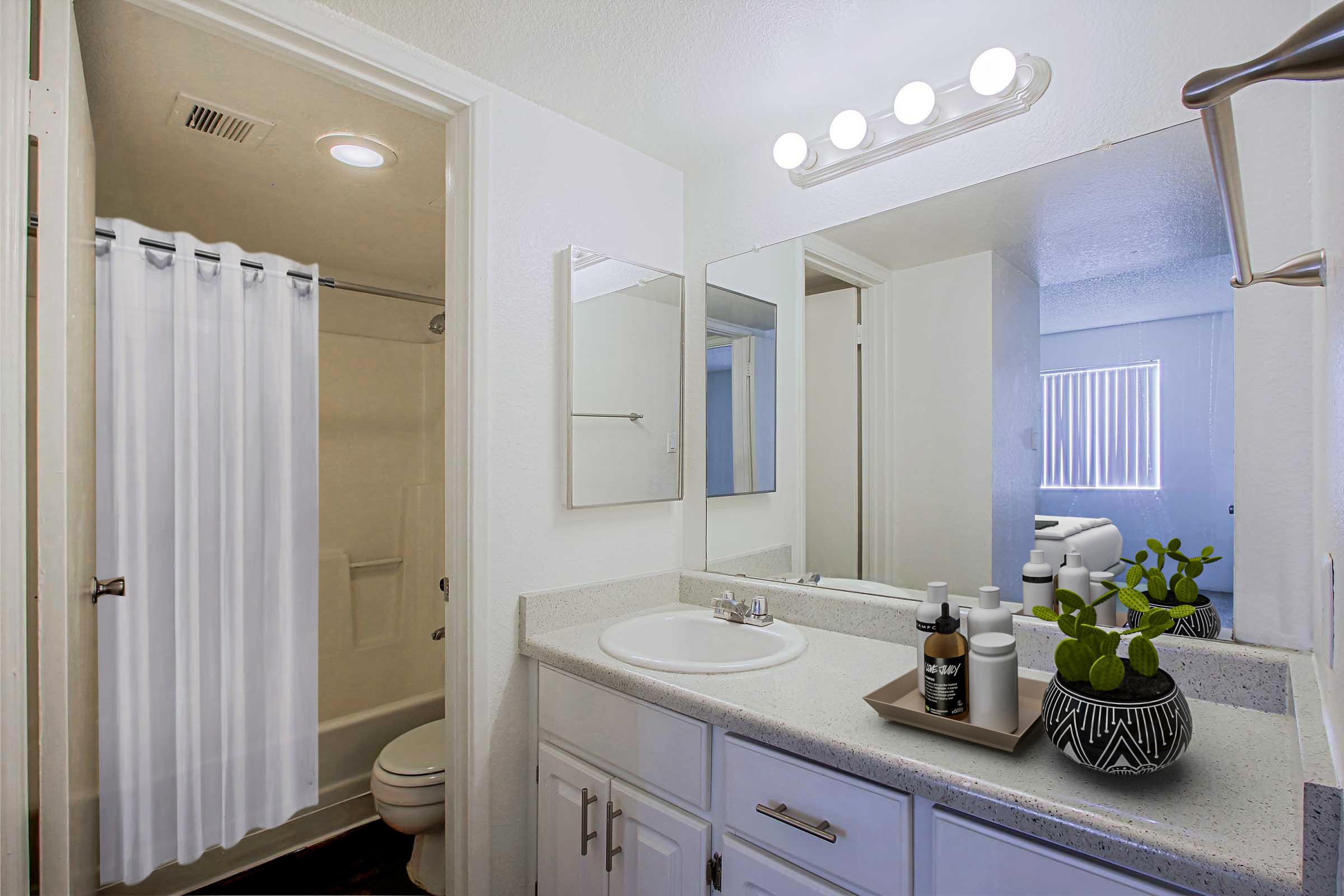 A modern bathroom featuring a shower with a white curtain, a clean vanity with a round mirror, and a sink. A tray with skincare products and a small potted plant is placed on the countertop. The walls are painted white, and there is a window providing natural light.