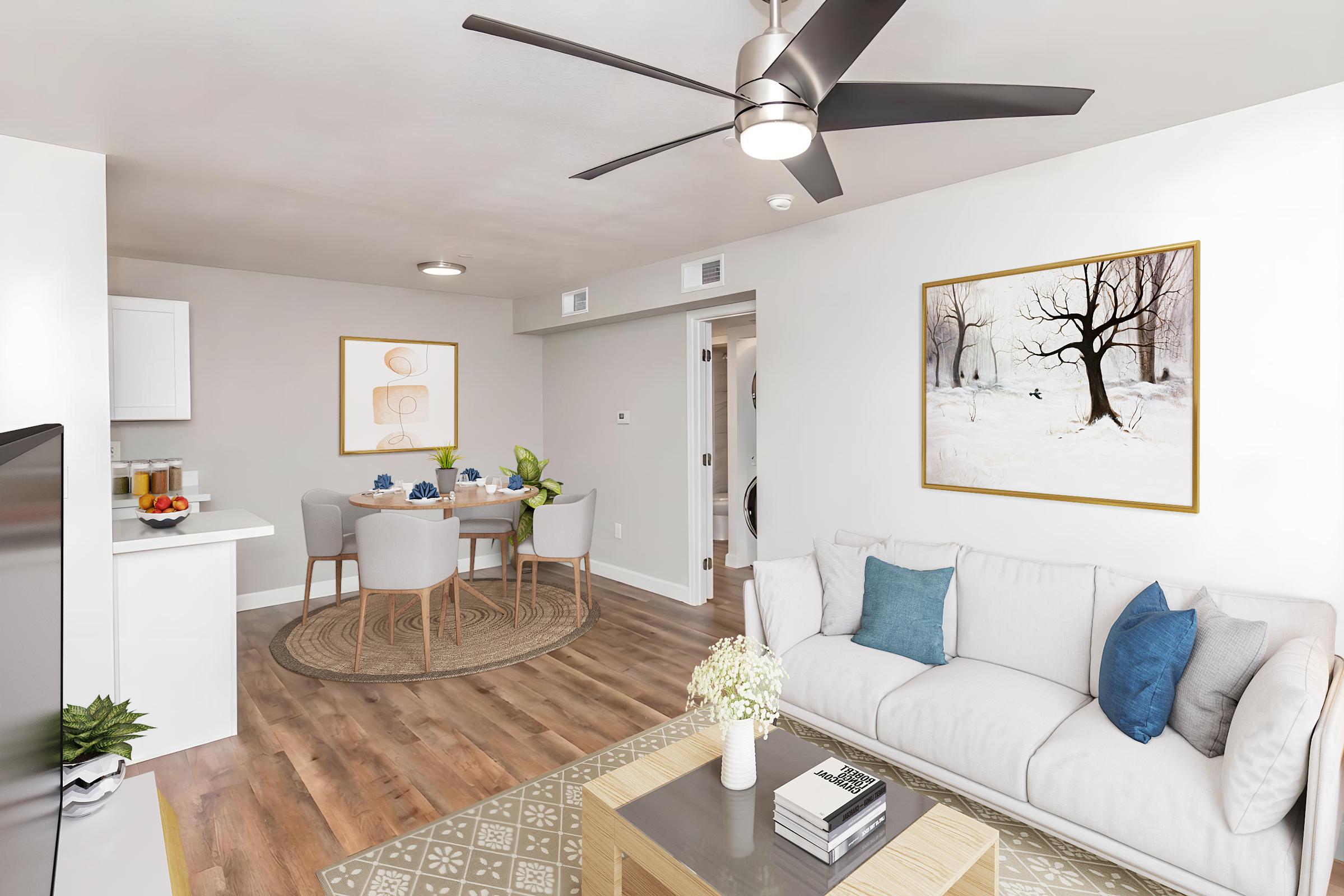 A modern living room featuring a light-colored sofa with blue cushions, a round coffee table, and a large TV. The space has a ceiling fan and a contemporary artwork on the wall. In the background, there's a dining area with a round table and four chairs, decorated with a vase and fruit. The overall decor is bright and welcoming.
