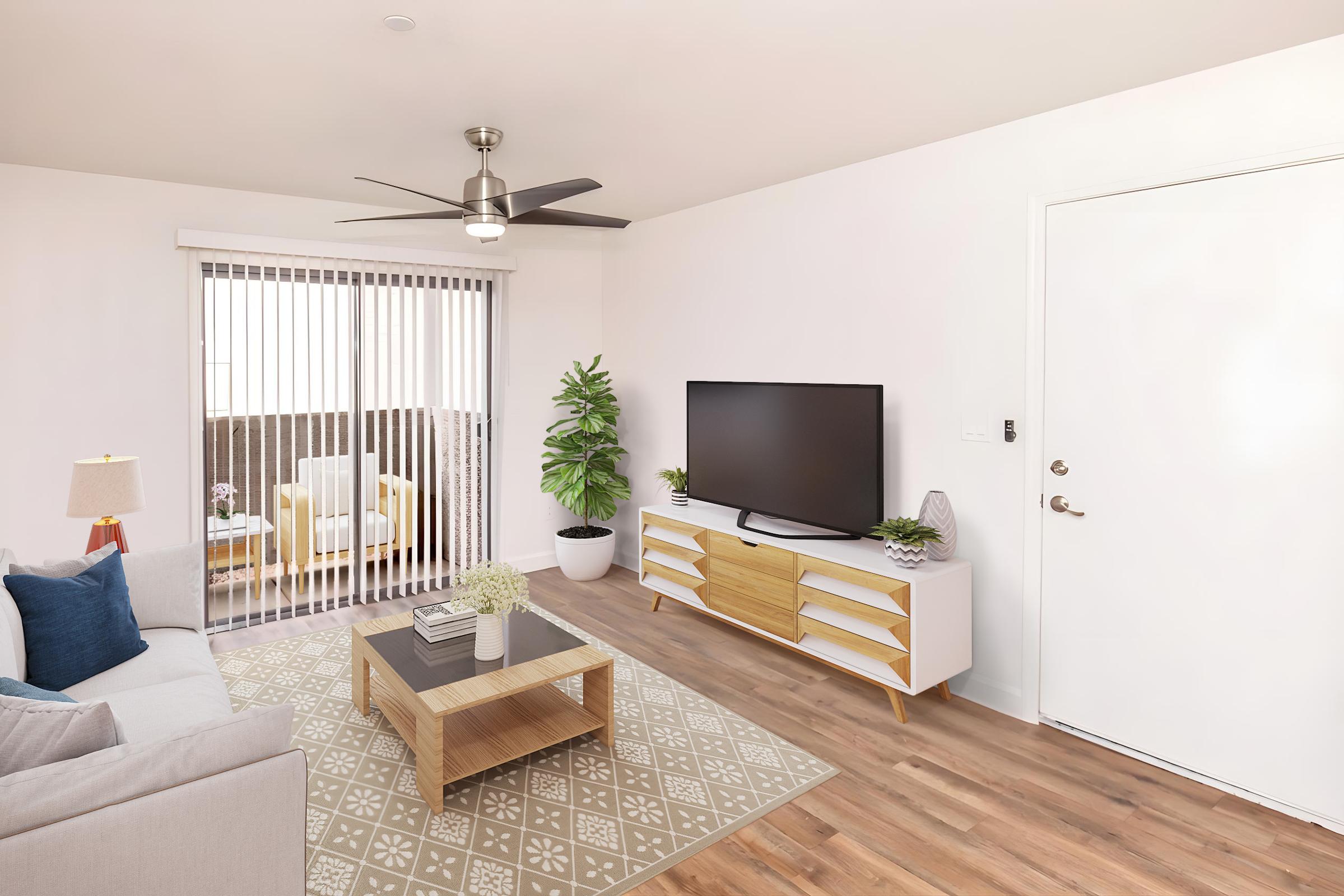 A modern living room featuring a light gray sofa, a wooden coffee table on a patterned area rug, and a large flat-screen TV on a white media console. There’s a potted plant beside the TV, a ceiling fan above, and sliding glass doors with vertical blinds leading to a balcony. A welcoming entry door is visible on the right.