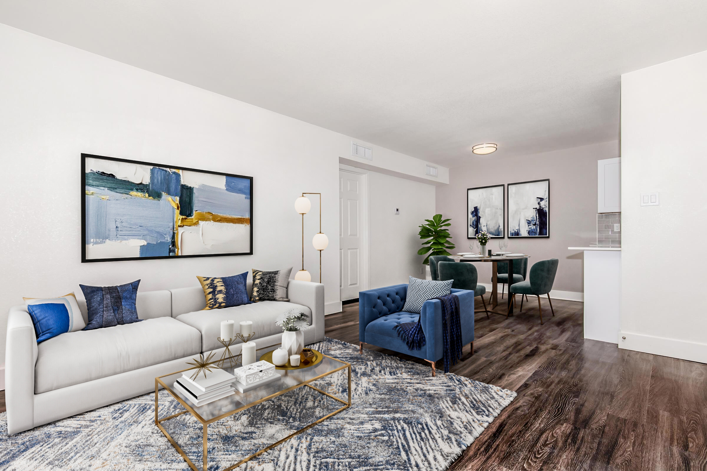 A modern living room featuring a white sofa with decorative pillows, a blue accent chair, and a gold coffee table. The space is adorned with abstract artwork on the walls, a plush area rug, and a green plant. In the background, there is a dining area with a small table and chairs.