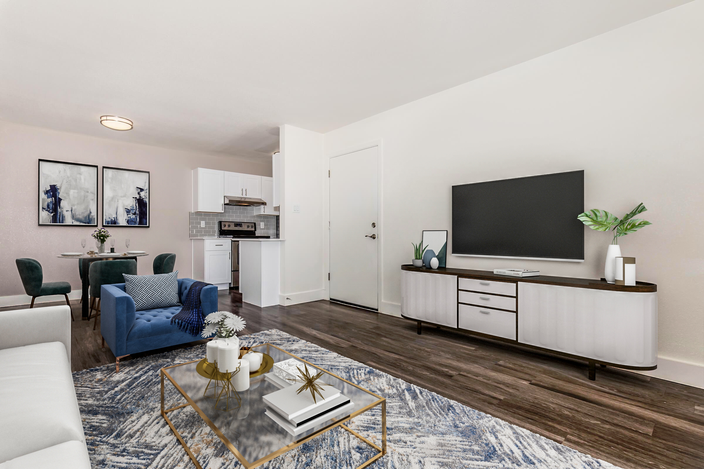 A modern living room and kitchen area featuring a blue armchair, a white sofa, and a coffee table with decorative items. There's a large TV on a wooden unit against the wall and a dining setup with green chairs in the background. The space is bright and contemporary, showcasing stylish decor and wooden flooring.