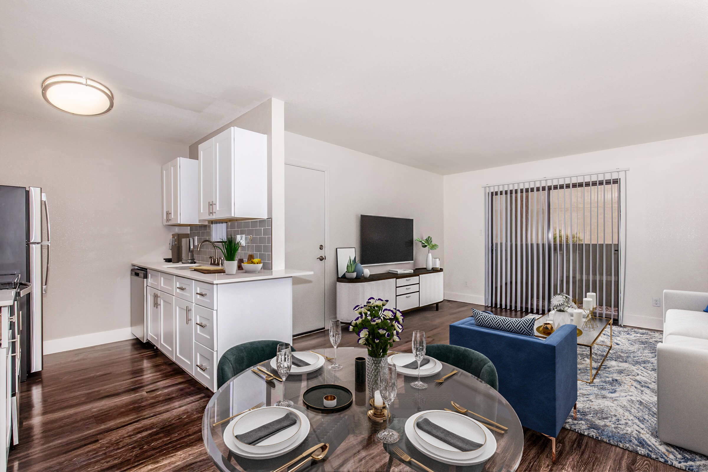 A modern living area featuring a dining table set for two, a cozy blue sofa, and a stylish coffee table. The kitchen is visible with white cabinets and stainless steel appliances. A television sits on a white console, and large windows with blinds allow natural light in, enhancing the bright and inviting atmosphere.