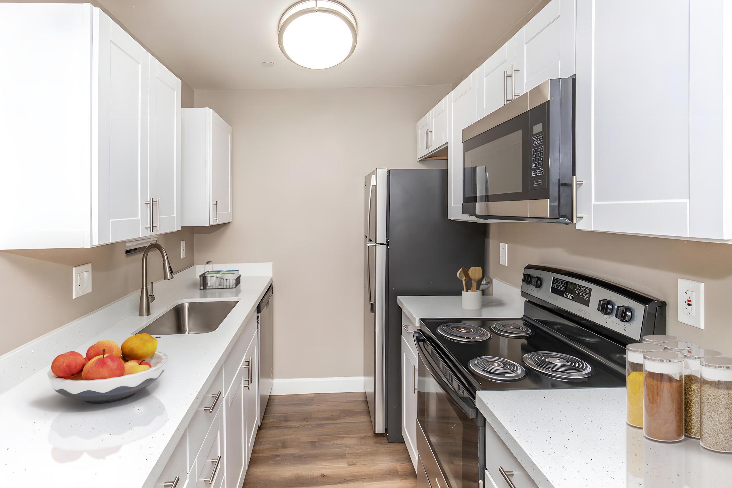 Modern kitchen with white cabinets, stainless steel appliances, and a dark countertop. Features include a double sink, microwave, and stove. A bowl of apples is on the counter, and several jars of spices sit neatly arranged. The space is well-lit with a round ceiling light.