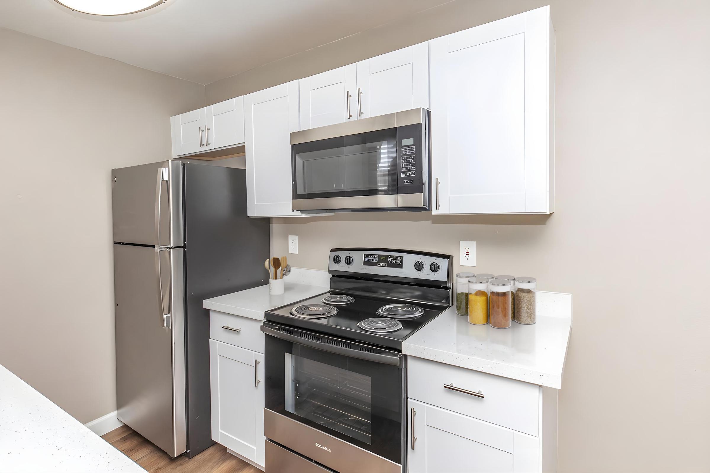 Modern kitchen featuring a stainless steel refrigerator, microwave above a sleek black stove, and white cabinetry. The countertop is white and has several jars of spices displayed. The walls are painted a neutral tone, contributing to a bright and airy atmosphere.