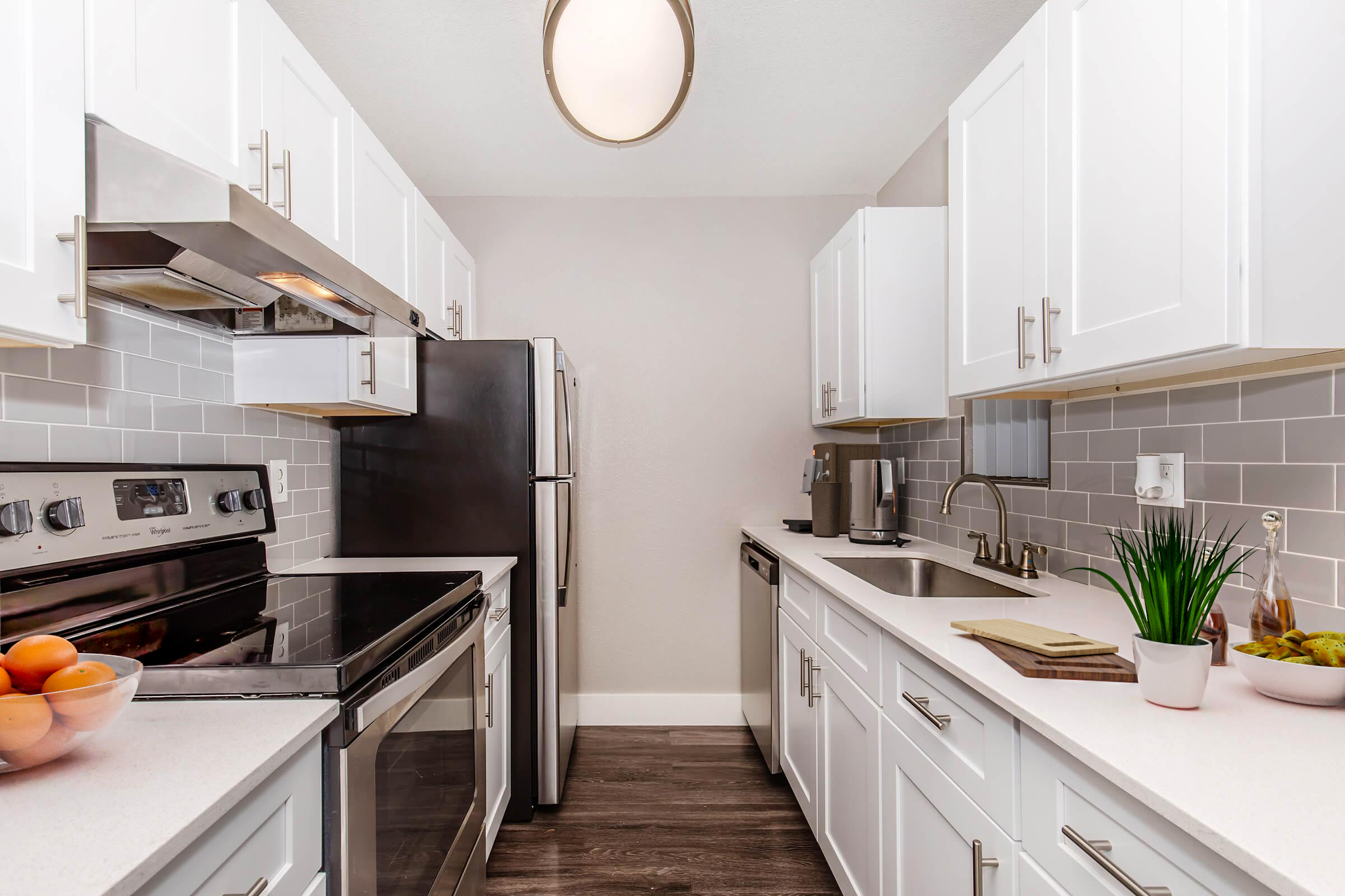 Modern kitchen featuring white cabinets, stainless steel appliances, and a gray tiled backsplash. The space includes an electric stove, oven, refrigerator, and sink, with a bowl of oranges on the counter. Soft lighting from a ceiling fixture enhances the clean, contemporary design.
