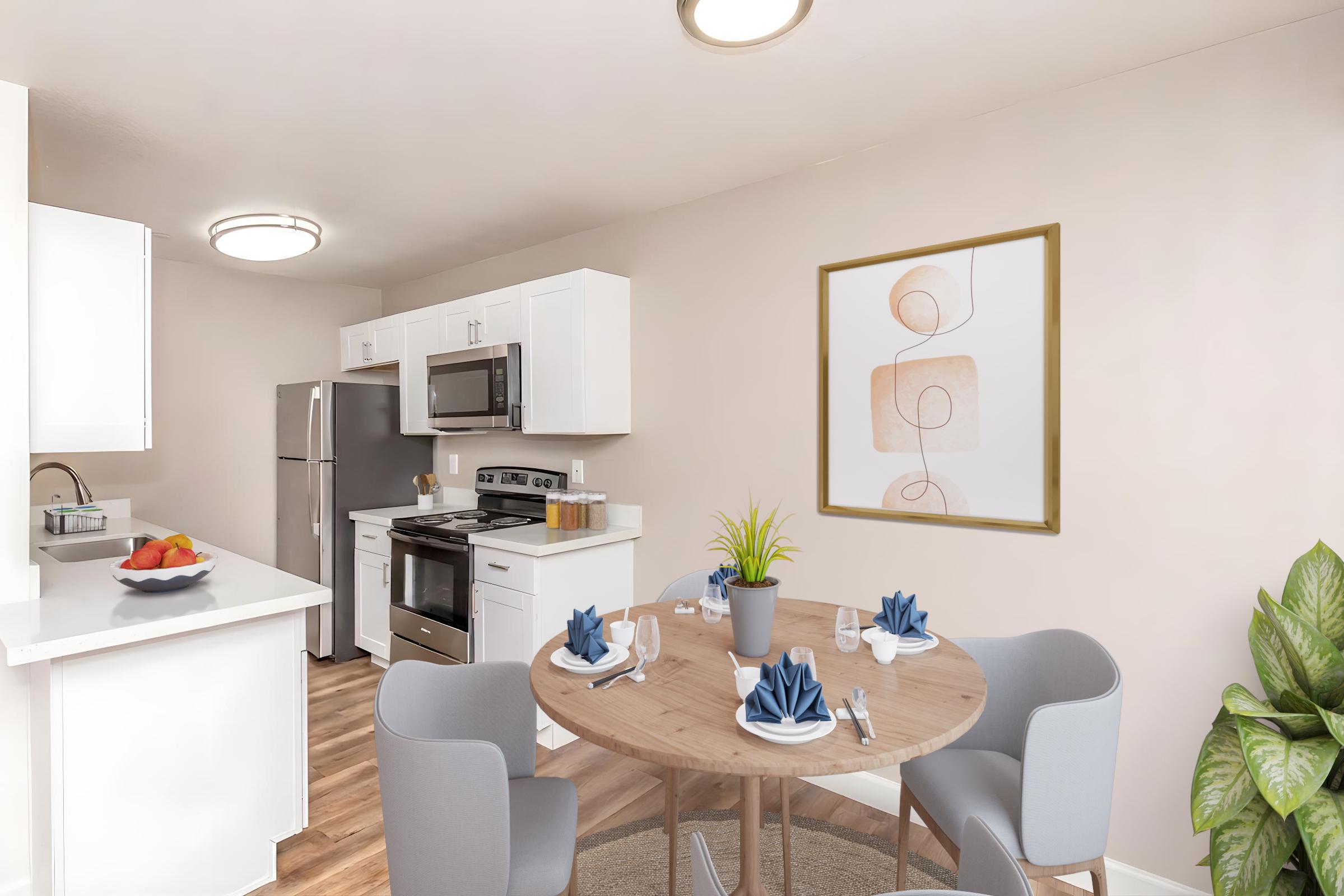 Modern kitchen and dining area featuring a round table set with blue napkins, a small plant centerpiece, and a plate of fruit. The kitchen has white cabinets, stainless steel appliances, and a light-colored wall with a framed abstract art piece. Natural light enhances the inviting atmosphere.