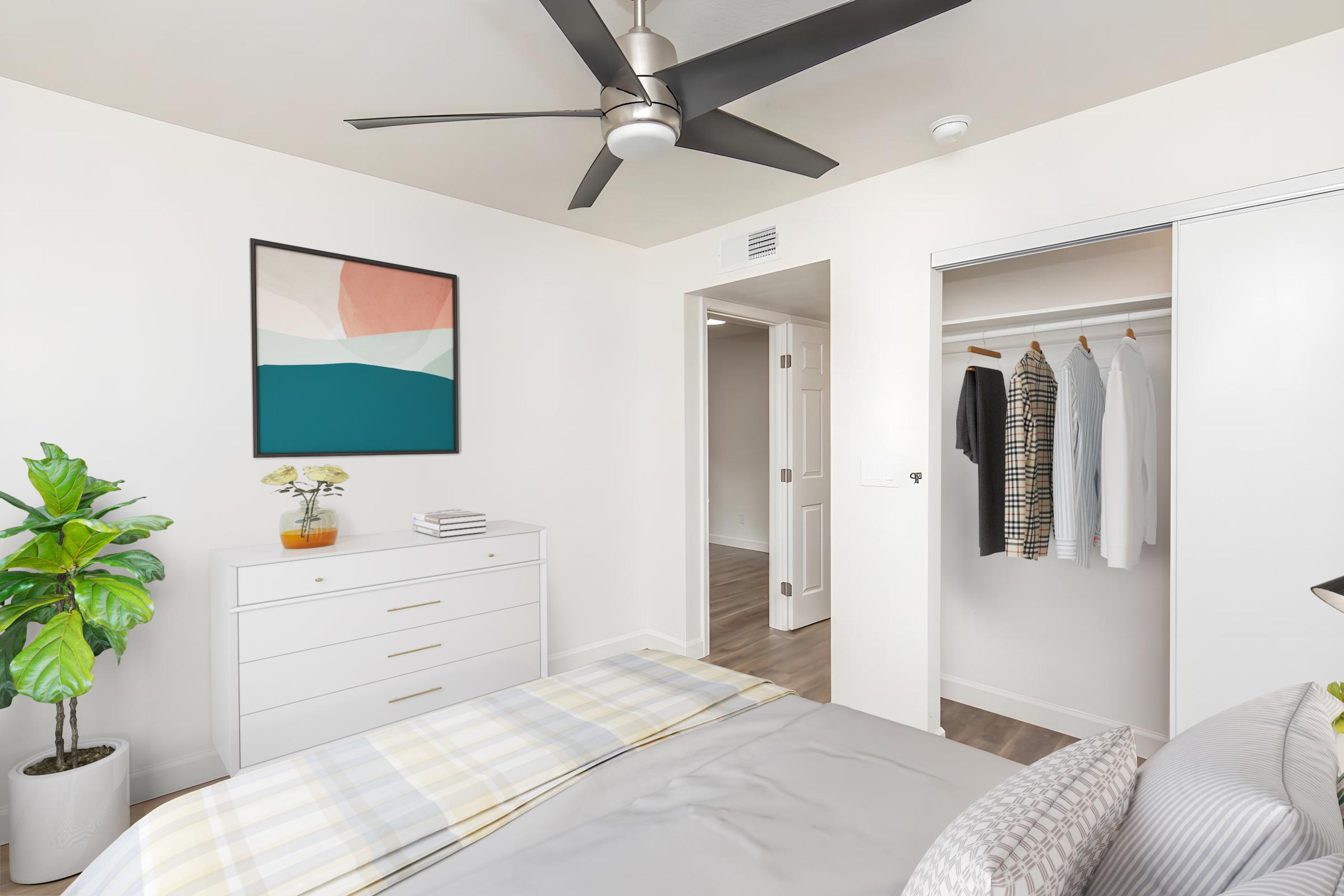 A bright and modern bedroom featuring a white dresser with a plant, a soft gray bedspread with a plaid pattern, a colorful abstract artwork on the wall, and a closet with neatly hung clothes. The space is well-lit, with a ceiling fan and a door leading to another room.