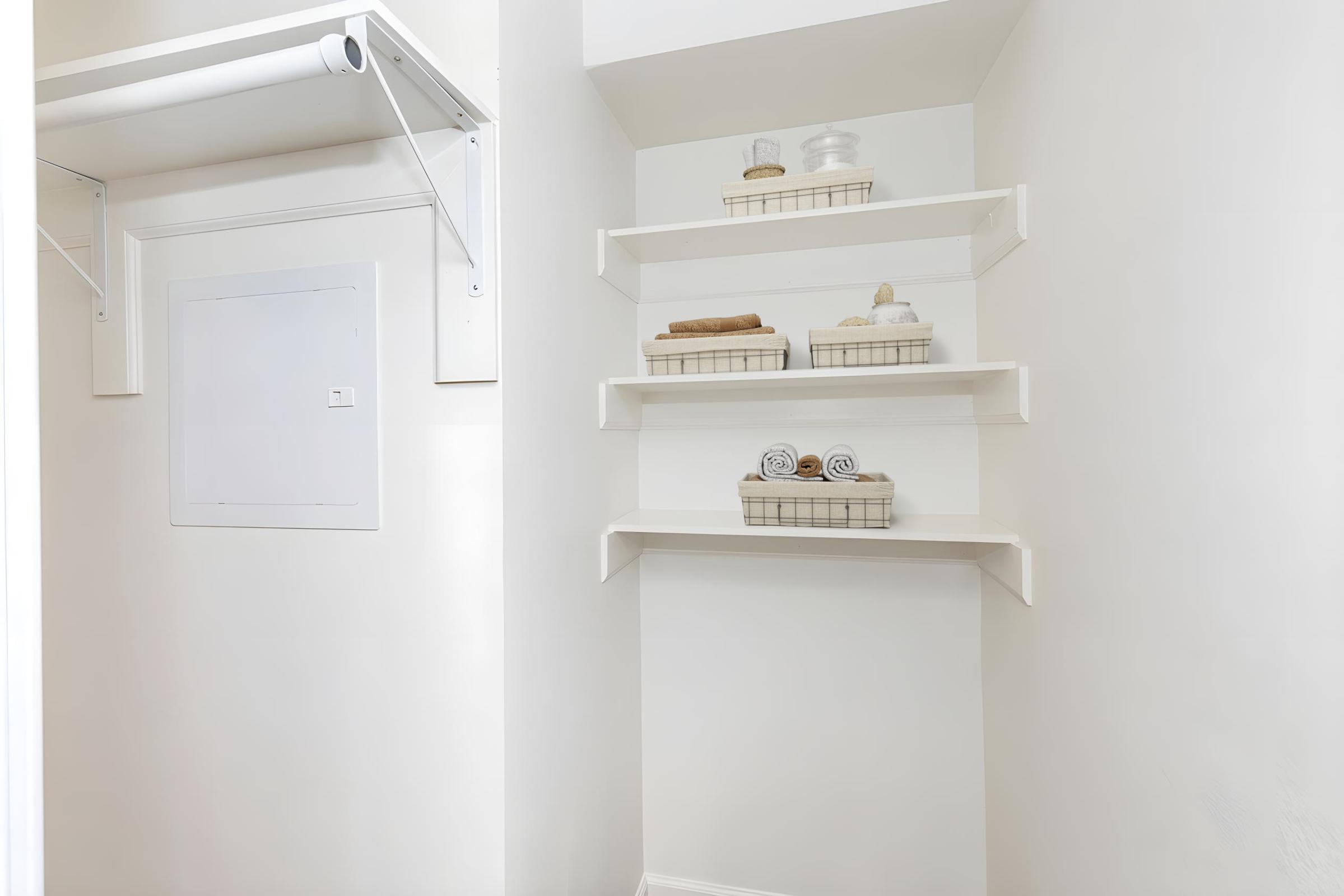 A clean, organized utility closet featuring white walls and shelves. The shelves display neatly folded towels and small storage baskets, while a wall-mounted rod is available for hanging items. A small door is visible on the wall, likely leading to additional storage or utilities.