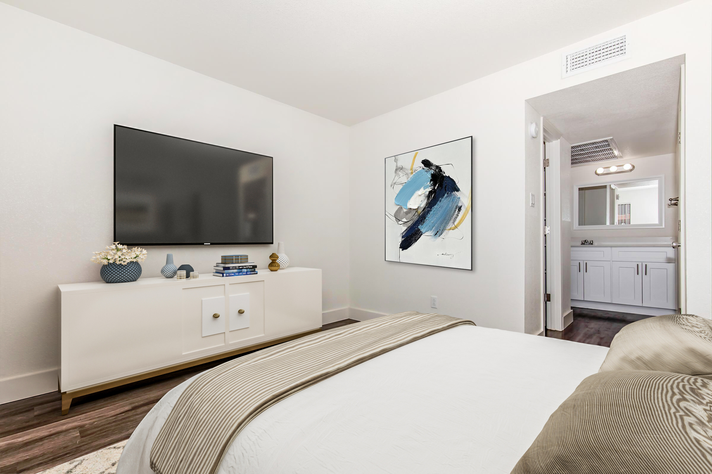 A modern bedroom featuring a white bed with a decorative beige blanket, a flat-screen TV mounted on the wall, and a stylish white dresser. An abstract painting with blue and black accents hangs beside the TV. A doorway leads to a bathroom area, showcasing bright lighting and contemporary design elements.