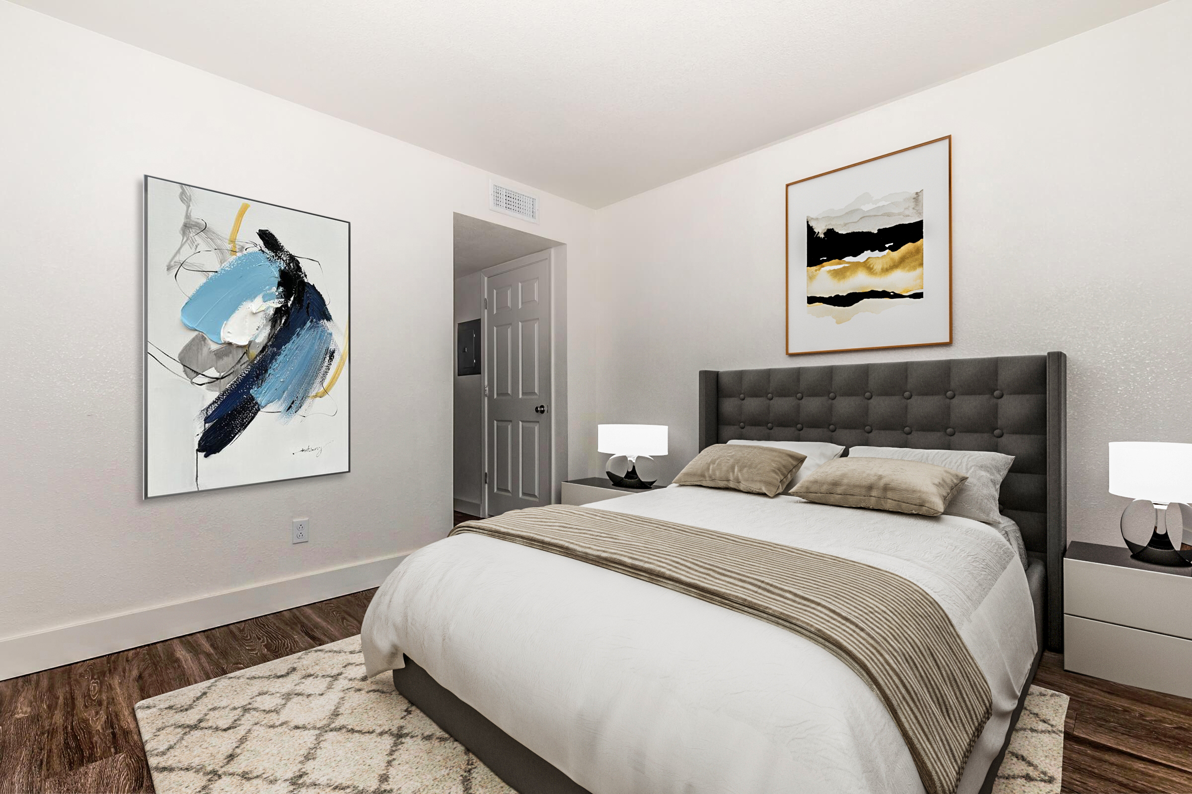 A modern bedroom featuring a gray tufted headboard with beige bedding, two bedside lamps, and artwork on the walls. The room has light-colored walls, hardwood flooring, and a neutral-toned area rug, creating a cozy and inviting atmosphere. A door is visible in the background.