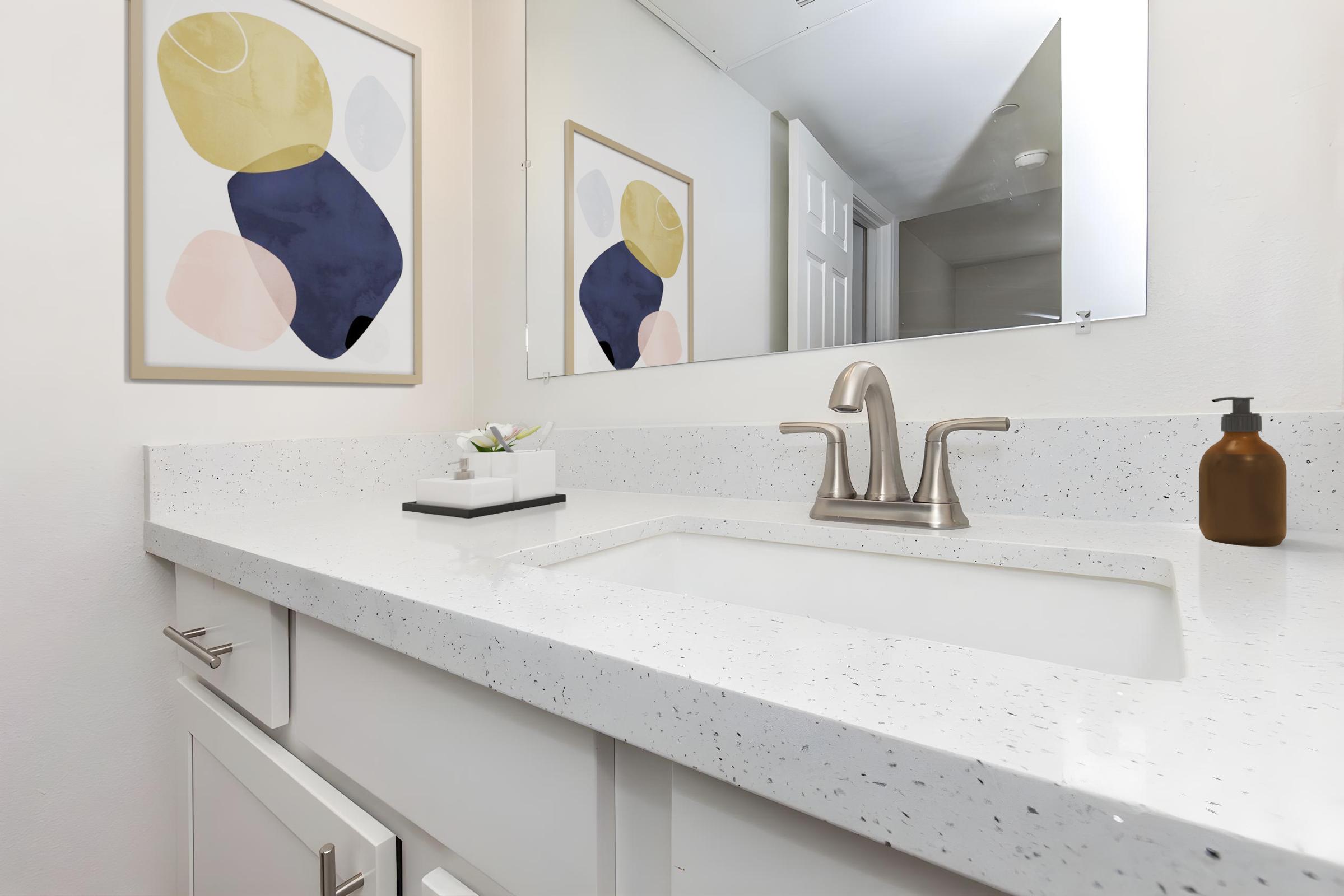 A modern bathroom vanity featuring a white countertop with a sink, a stylish faucet, and a decorative soap dispenser. Above the sink, there's a mirror reflecting the room, complemented by an abstract art piece on the wall, showcasing soft colors in a contemporary design.
