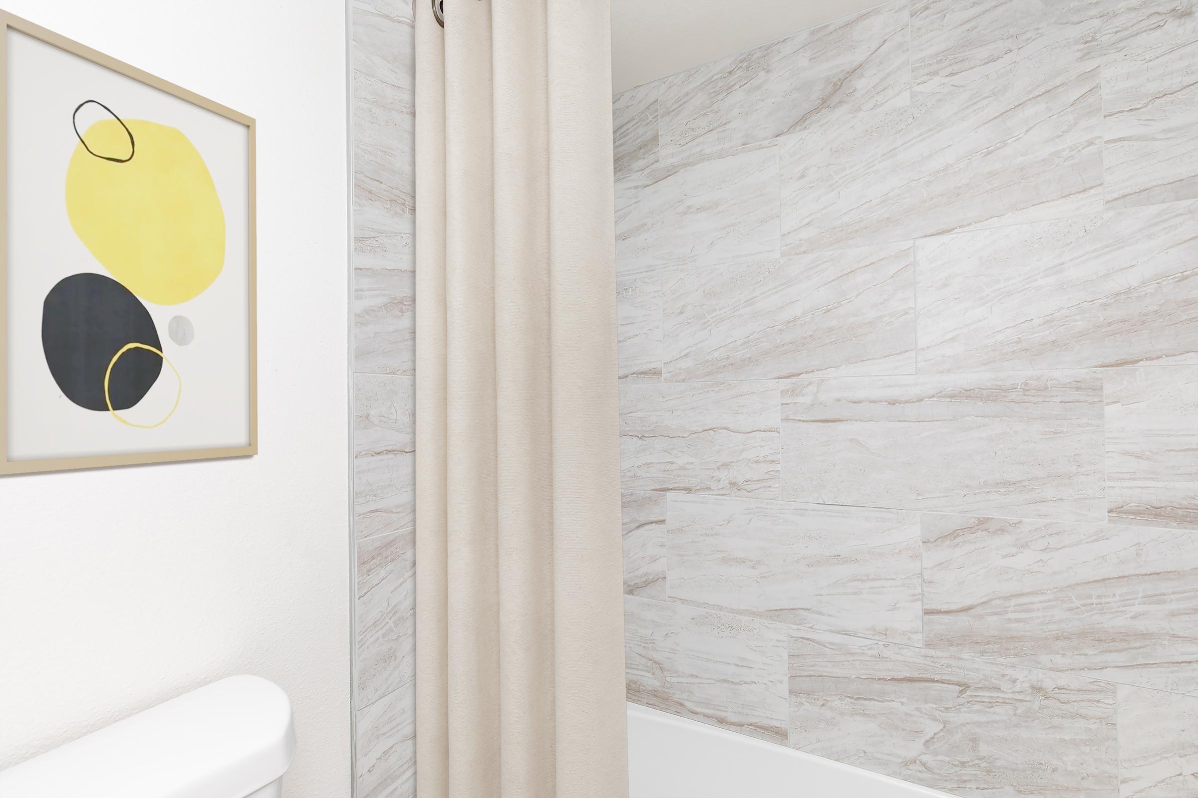 A bathroom scene featuring a light-colored tile wall, a shower curtain in a neutral tone, and a framed abstract artwork with yellow and black shapes on the wall. The bathtub is visible, and the overall aesthetic is modern and minimalistic.