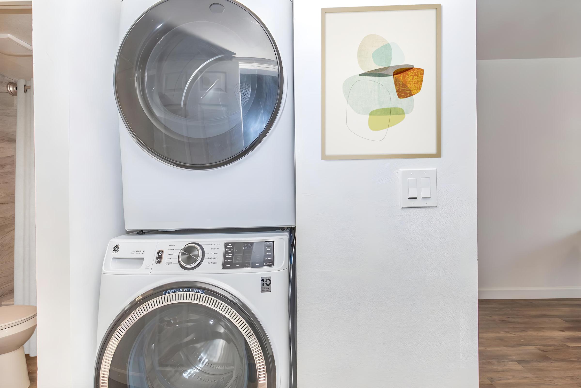 A stacked washer and dryer in a modern laundry space next to an abstract art piece in a frame. The walls are painted white, and a light-colored floor adds to the bright, clean ambiance. A glimpse of a bathroom is visible in the background.