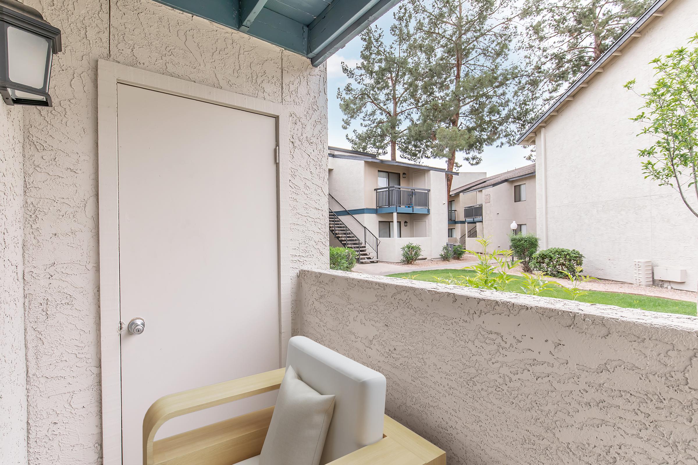 View from a balcony featuring a beige wall and a closed white door. A wooden chair with a cushion is positioned on the balcony, overlooking a green lawn and neighboring apartments with trees in the background. The scene is bright and inviting, suggesting a peaceful outdoor space.