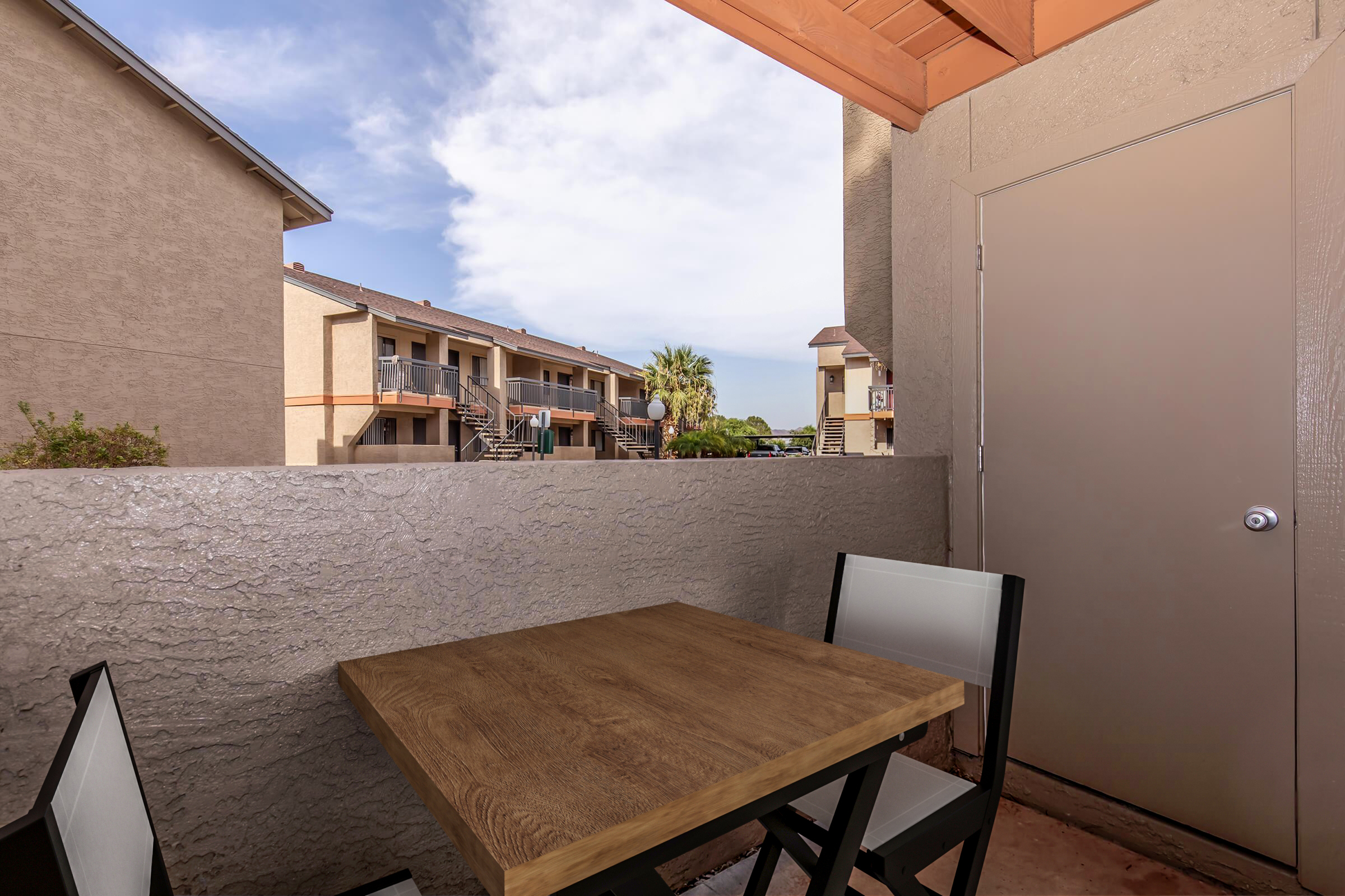 A small balcony view featuring a wooden table and two chairs. In the background, there are two residential buildings and a clear blue sky with scattered clouds. The space is well-lit, creating a welcoming outdoor atmosphere.