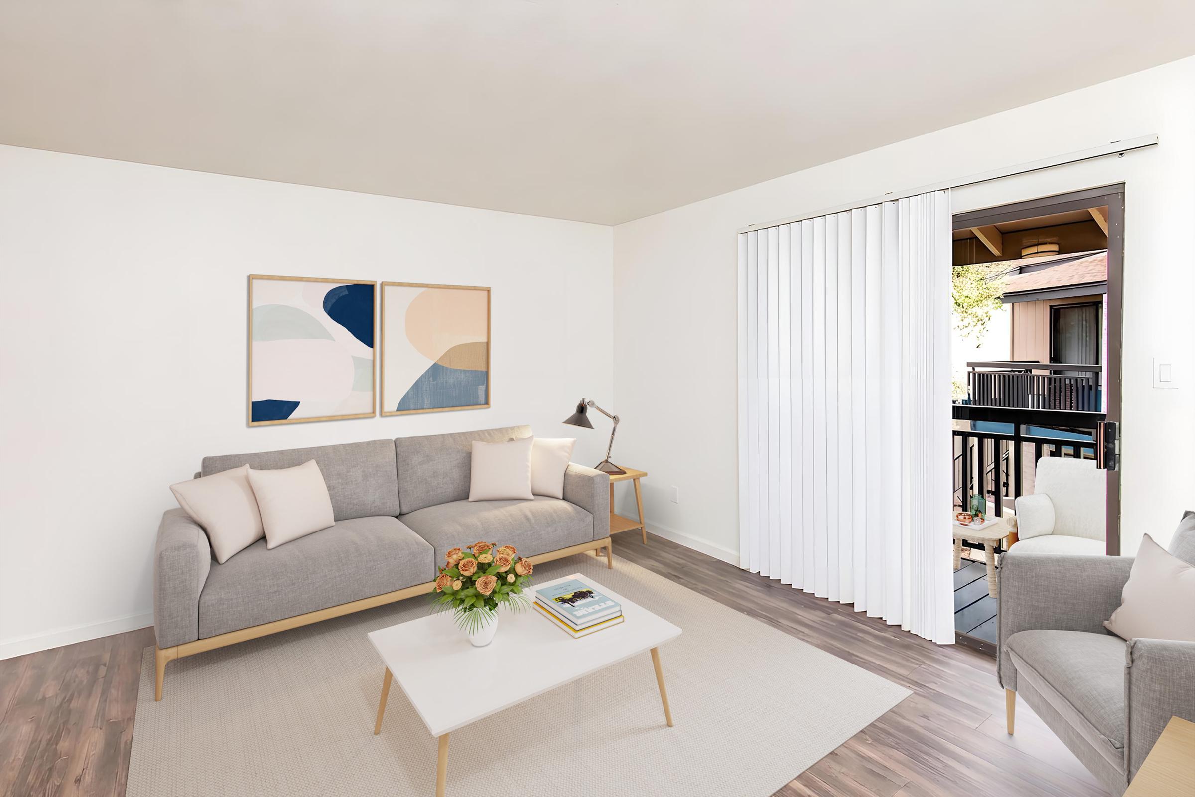 A modern living room featuring a light gray sofa with wooden legs, a minimalist coffee table, and a chair. Two abstract artworks are hung on the wall, and a vase of flowers sits on the table. A sliding door leads to an outdoor balcony, with sheer curtains partially drawn. The space has neutral tones and natural light.
