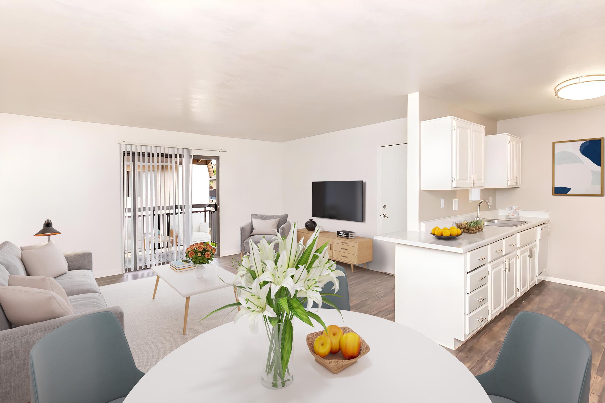 A modern living room with a light-colored theme featuring a gray sofa, a small coffee table with flowers, and a dining area with a round table and fruit. A kitchen with white cabinets is visible in the background, alongside a wall-mounted TV and a door leading to a balcony.