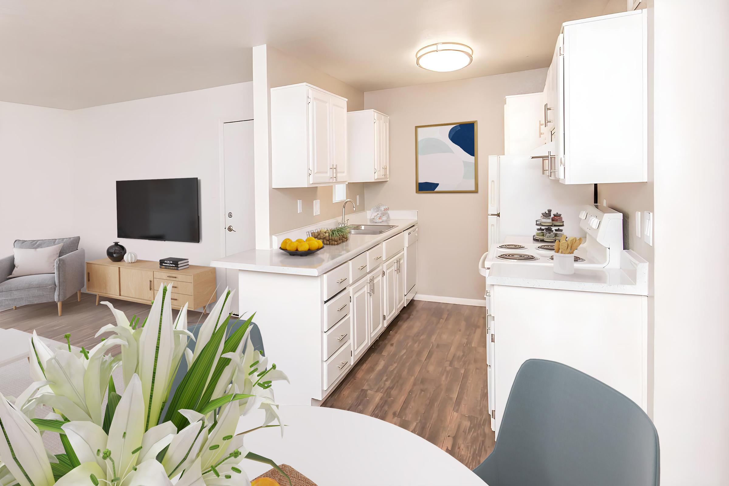 Bright and modern kitchen area featuring white cabinetry, a countertop with fresh fruit, and appliances. In the background, a cozy living room with a TV and a light gray sofa can be seen. A dining table with a floral centerpiece completes the inviting space.