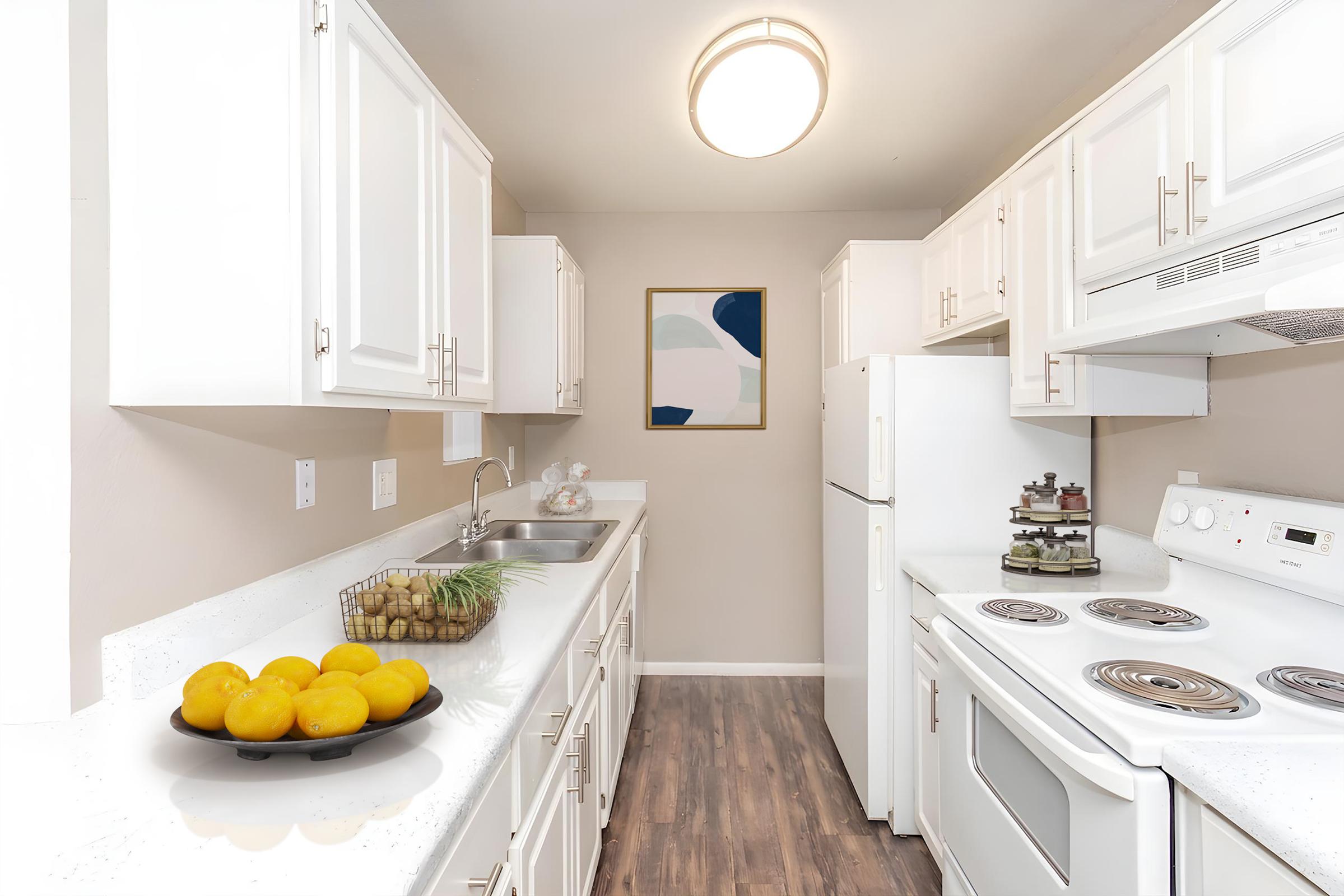 A modern kitchen featuring white cabinets and appliances, a sink on the left, and a stove on the right. A bowl of oranges is on the counter, and there's a decorative piece on the wall. The flooring is dark wood, creating a bright and inviting atmosphere.