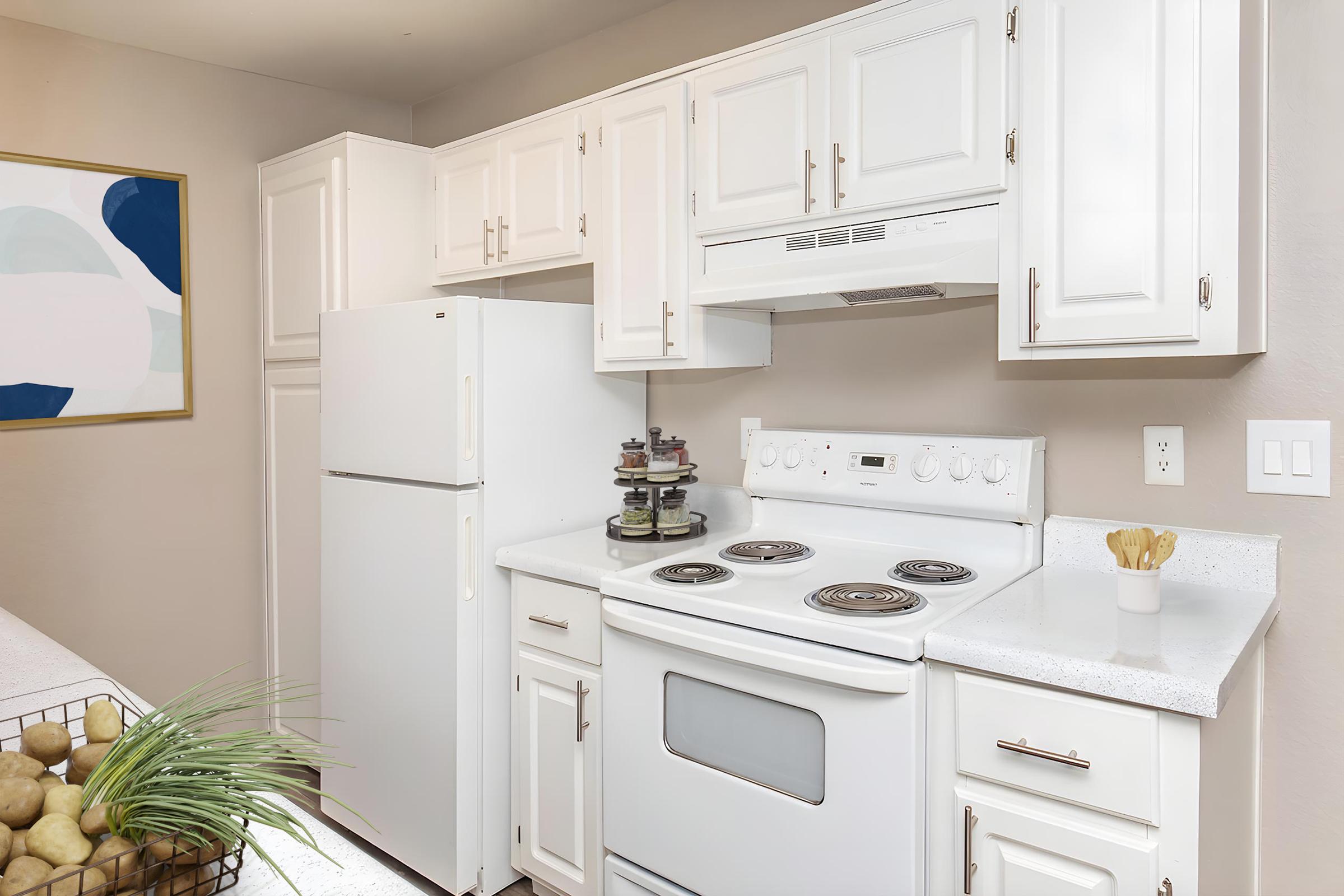 Bright kitchen featuring white cabinetry, a white refrigerator, and a stove with an overhead exhaust. The countertop is light-colored with a bowl of decorative fruits and a small plant. A piece of abstract art is hung on the wall, adding a splash of color to the space.