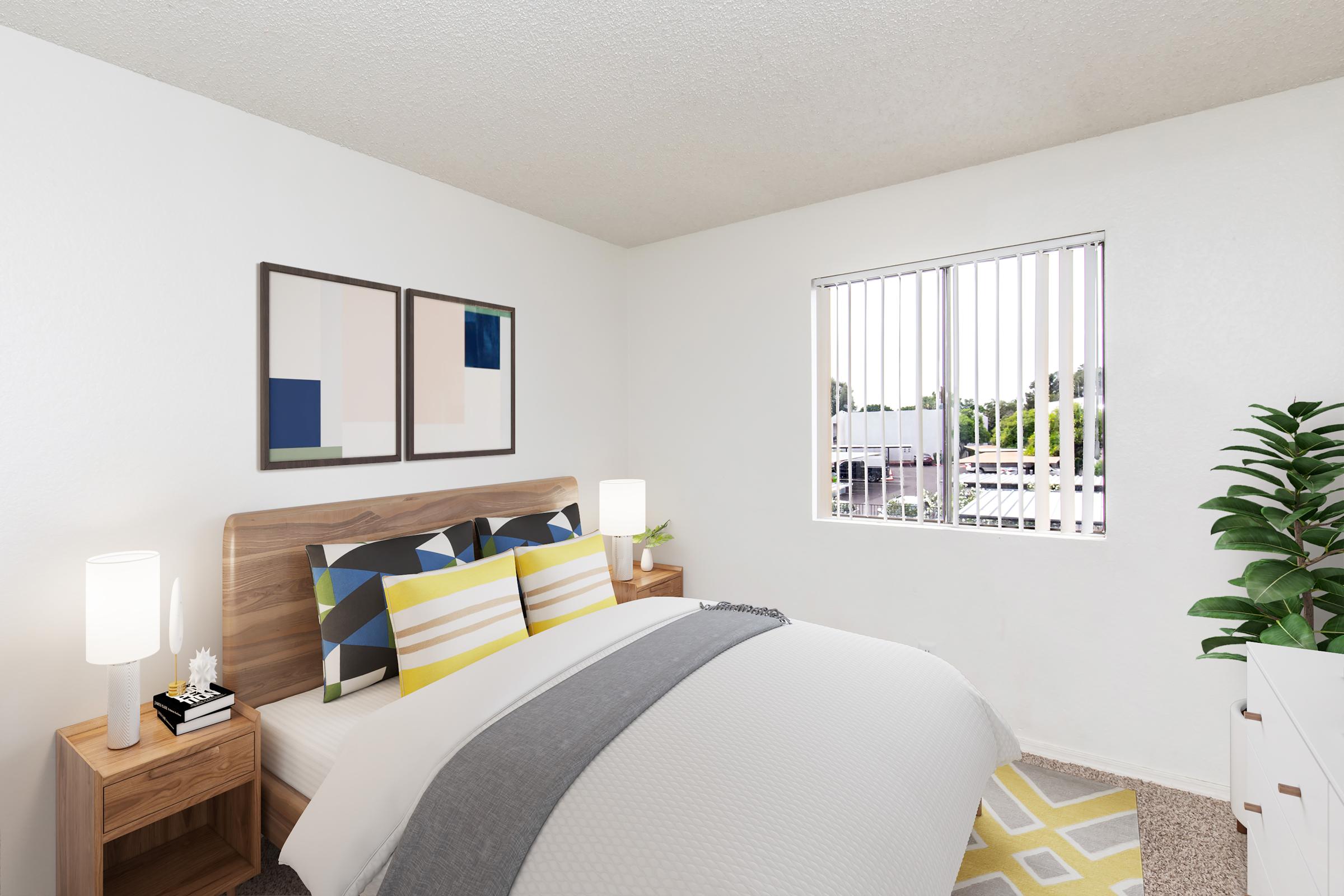 A cozy bedroom featuring a wooden bed with colorful bedding. Two bedside lamps illuminate framed abstract art on the wall. There’s a window with blinds allowing natural light, and a green potted plant adds a touch of nature. The room has a light color scheme and a patterned rug on the floor.