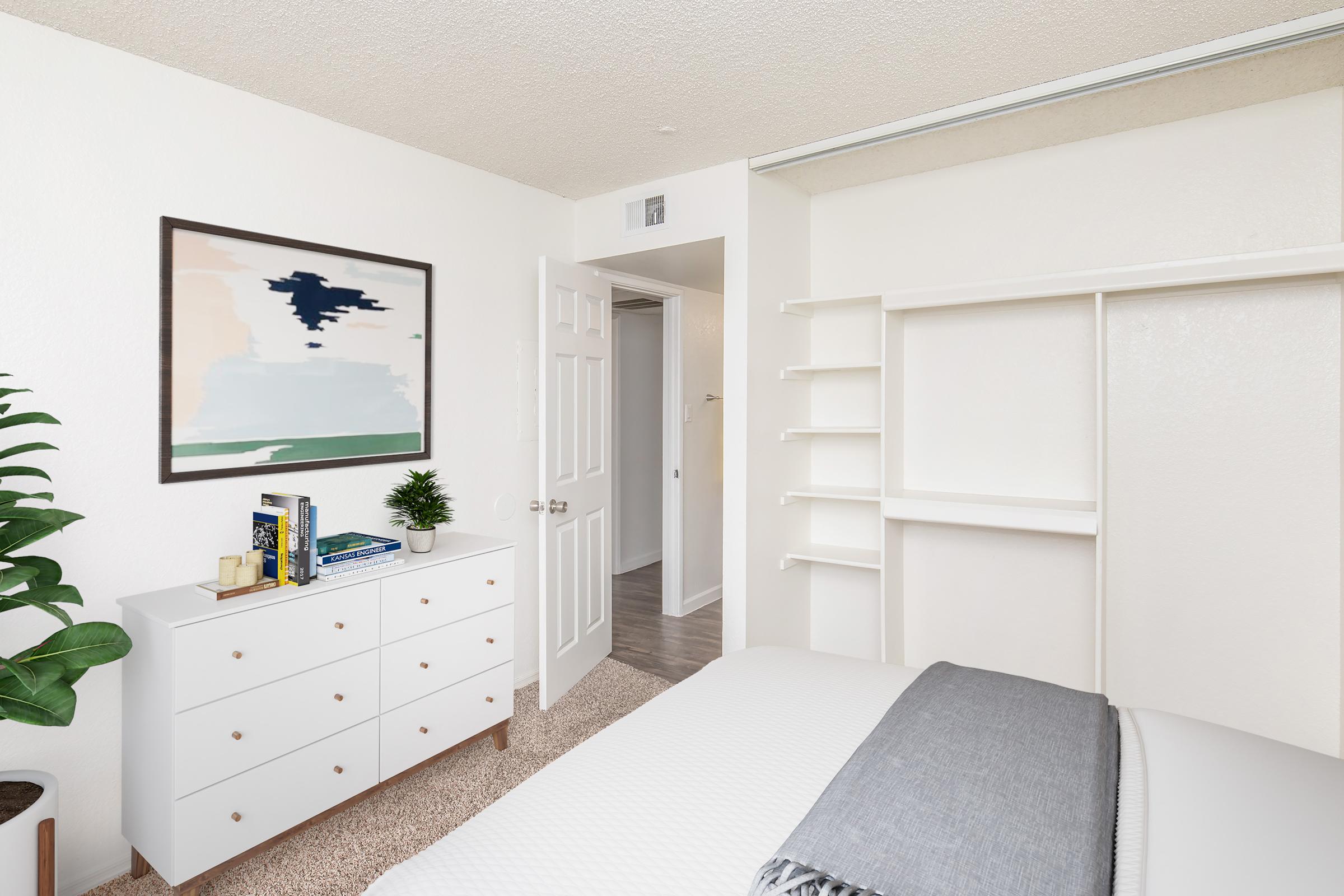 A cozy bedroom featuring a white dresser with books and a small potted plant, a simple bed with gray bedding, and an abstract painting on the wall. The room has light-colored walls, carpeted flooring, and a glimpse of a doorway leading to another space.