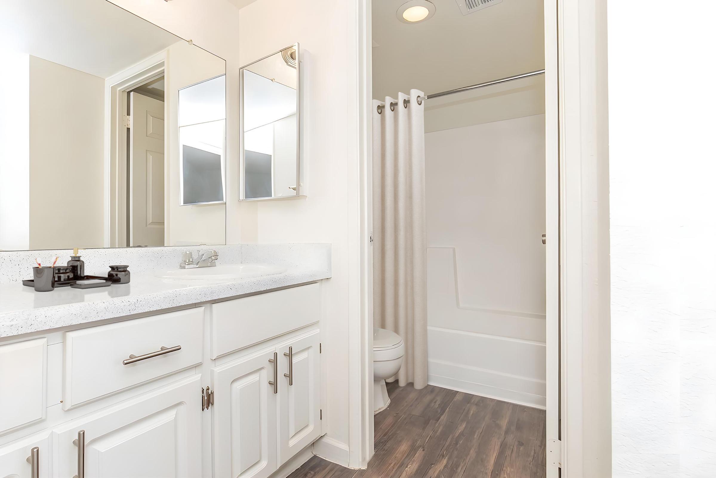 A bright and modern bathroom featuring a white vanity with a sink, twin mirrors above it, and a light-colored countertop. A shower area with a curtain is visible, along with a toilet. The flooring is a dark wood laminate, adding contrast to the overall light decor.