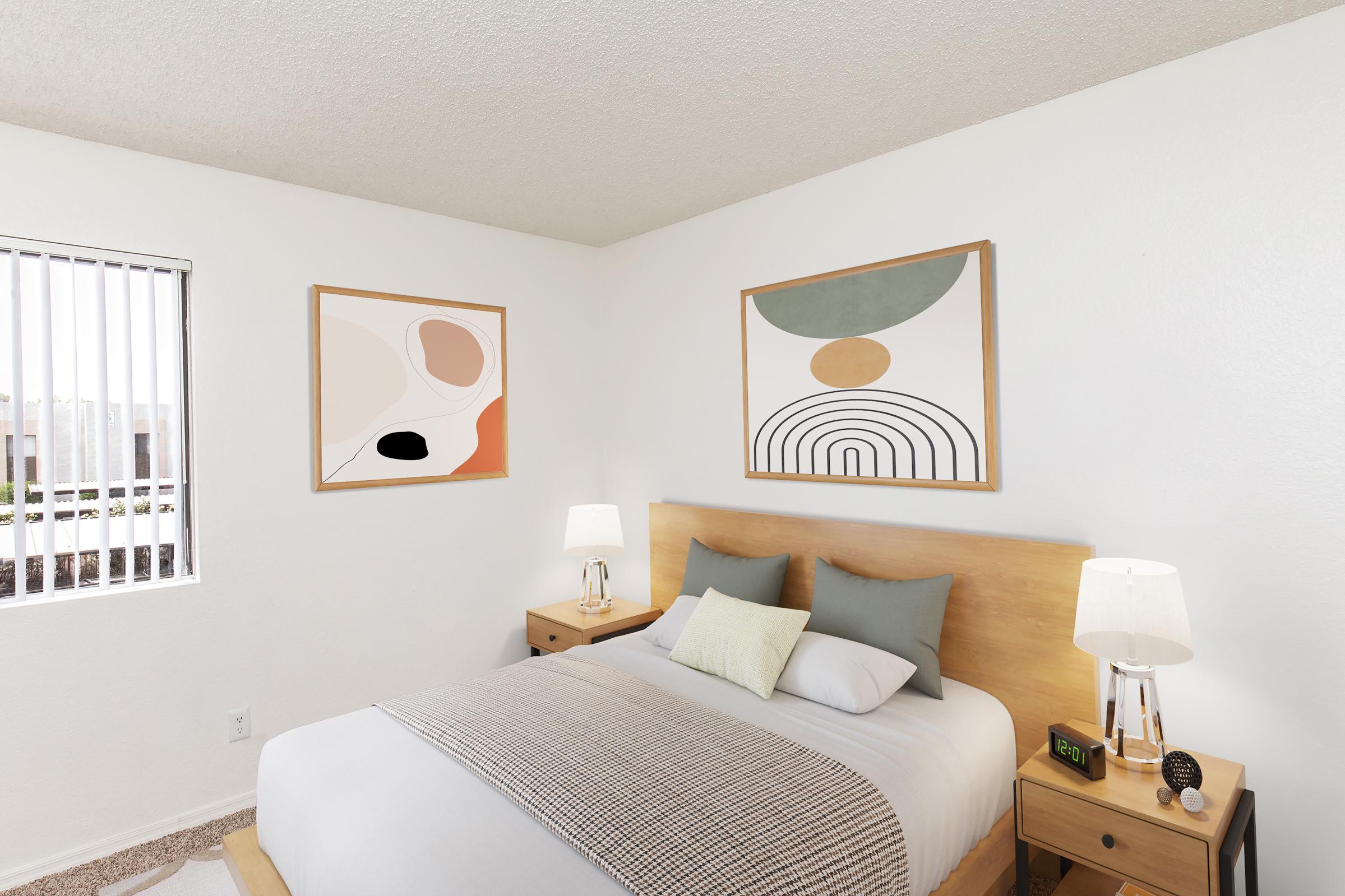 A cozy bedroom featuring a double bed with light bedding, accent pillows, and wooden nightstands with lamps. Two abstract art pieces hang on the wall, and there's a window with vertical blinds letting in natural light. The room has a warm and inviting atmosphere.