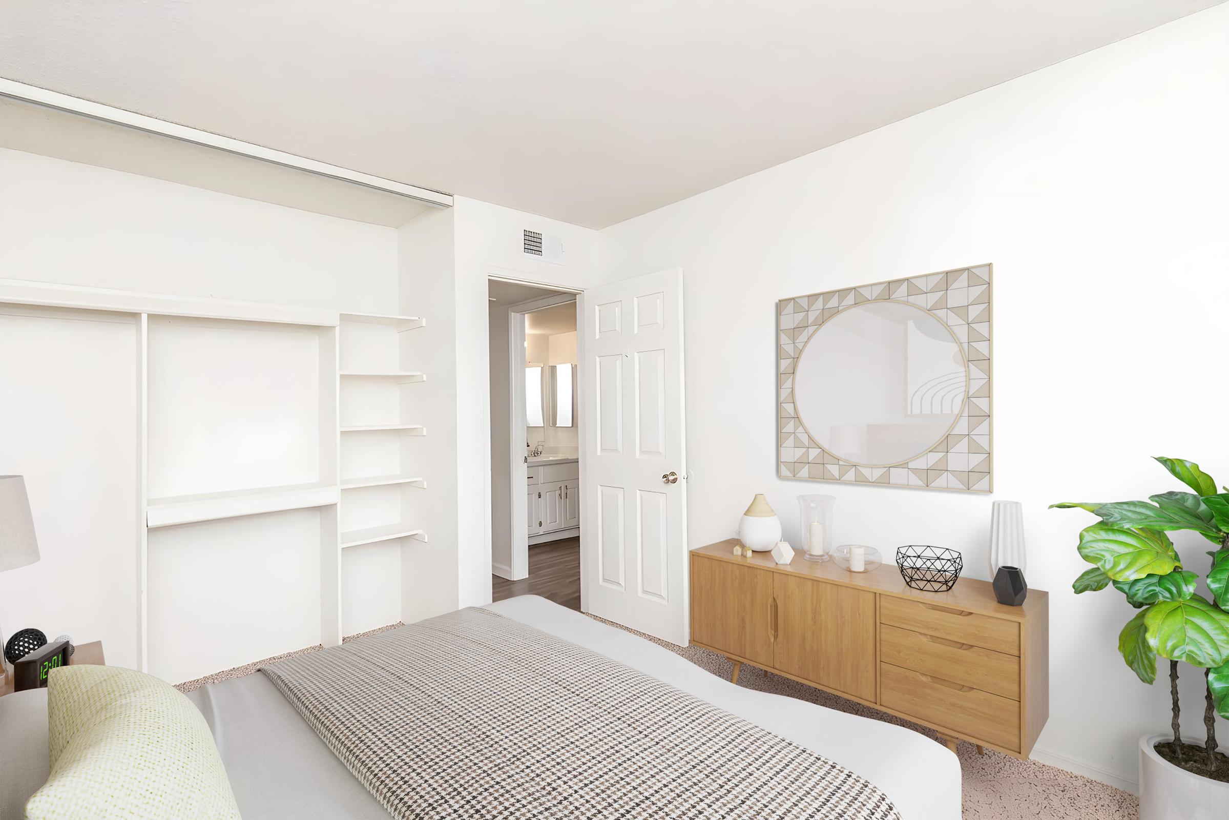 A modern bedroom featuring a large bed with a patterned blanket, a decorative mirror on the wall, and a wooden dresser with decorative items. There are open shelves and a potted plant, with a door leading to a bathroom in the background. The room has a bright, minimalist aesthetic.