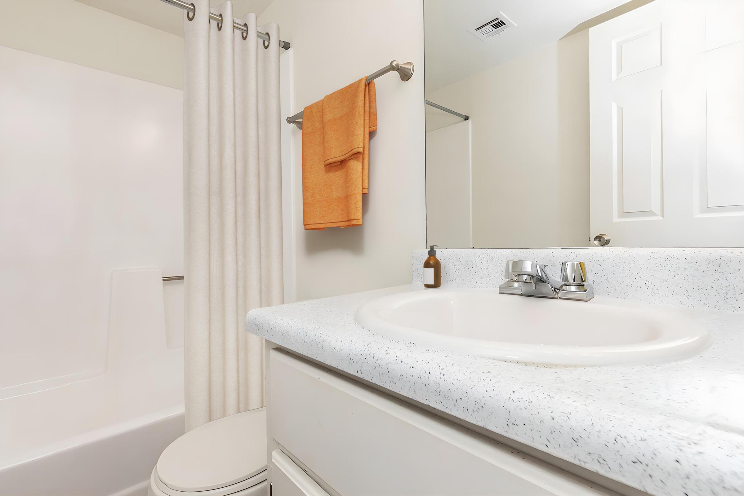 A clean and modern bathroom featuring a white countertop with a round sink, silver faucet, and soap dispenser. A shower area is visible with a light-colored curtain, and an orange towel hangs on the towel rack. The bathroom has neutral-colored walls and a white toilet.