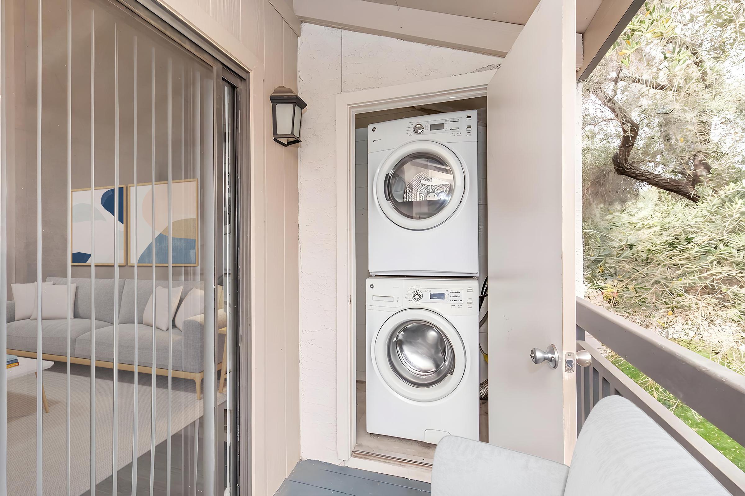A compact laundry area featuring a stacked washer and dryer inside a doorway, with a view of a cozy living space and greenery outside through the adjacent glass sliding door. The setting is bright and inviting.