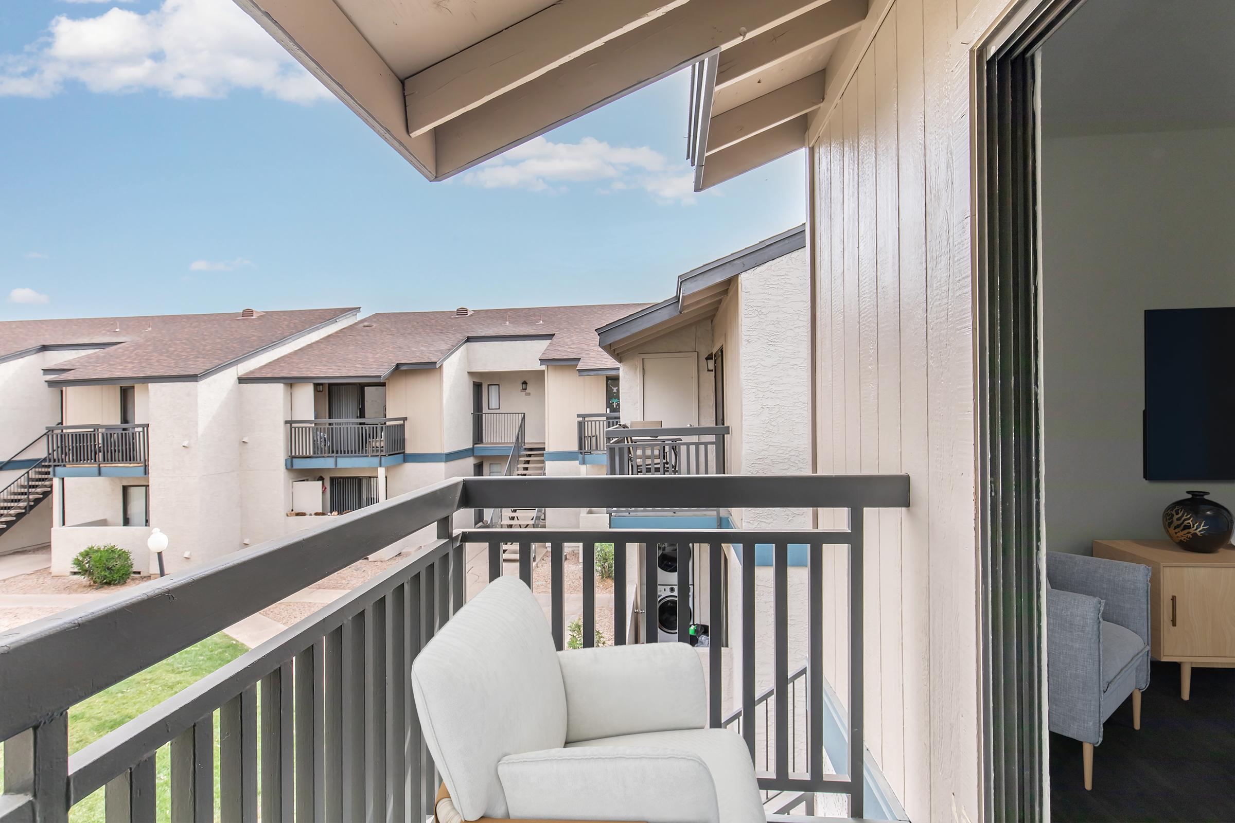 A balcony view of an apartment complex, featuring a comfortable white chair and a glimpse of neighboring units. The scene includes a clear blue sky and well-maintained landscaping, creating a tranquil outdoor space.