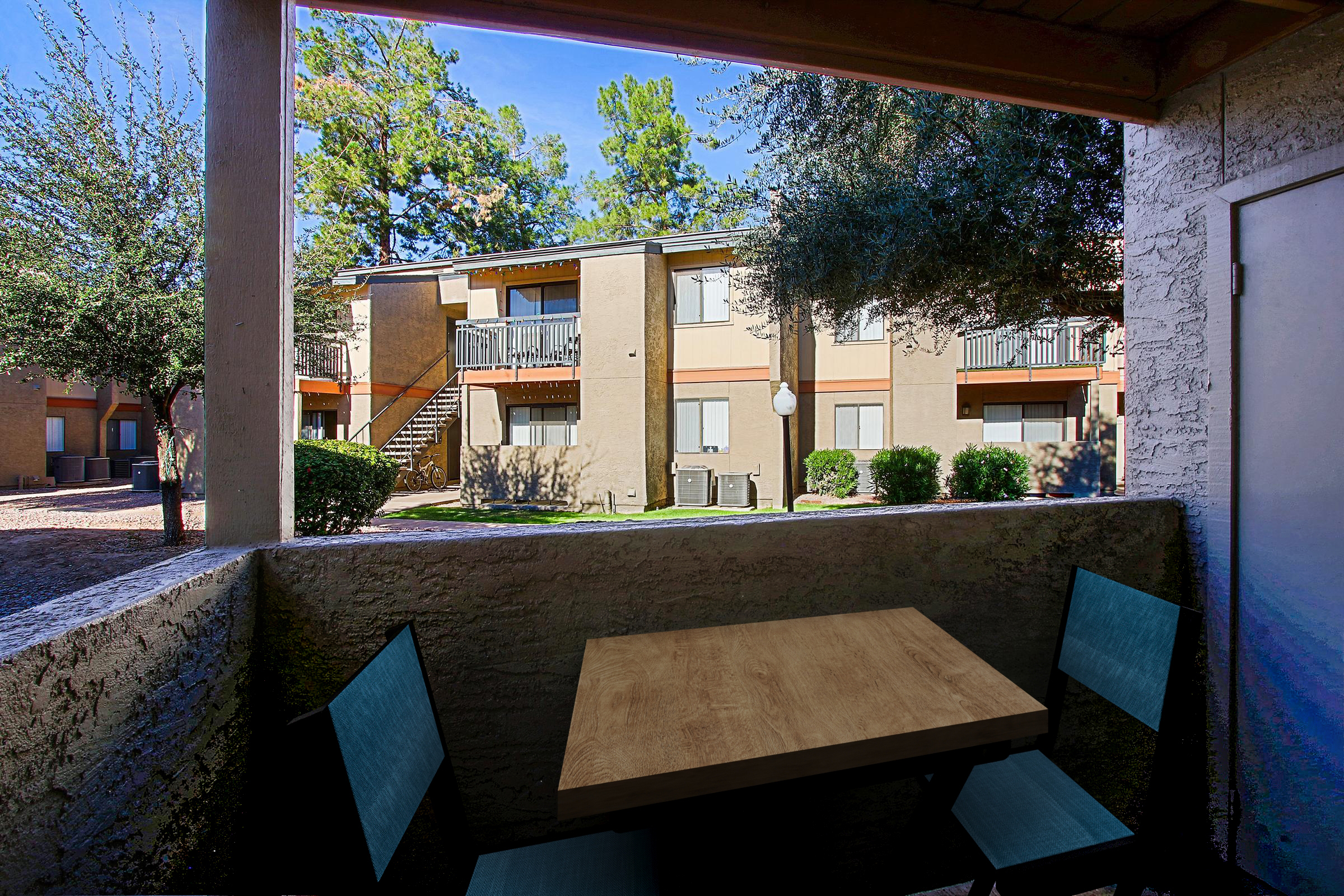 View from a porch showcasing a small wooden table with two blue chairs, overlooking a landscaped area and apartment buildings with balconies. Trees are visible in the background, and the sky is clear and sunny, creating a welcoming outdoor atmosphere.