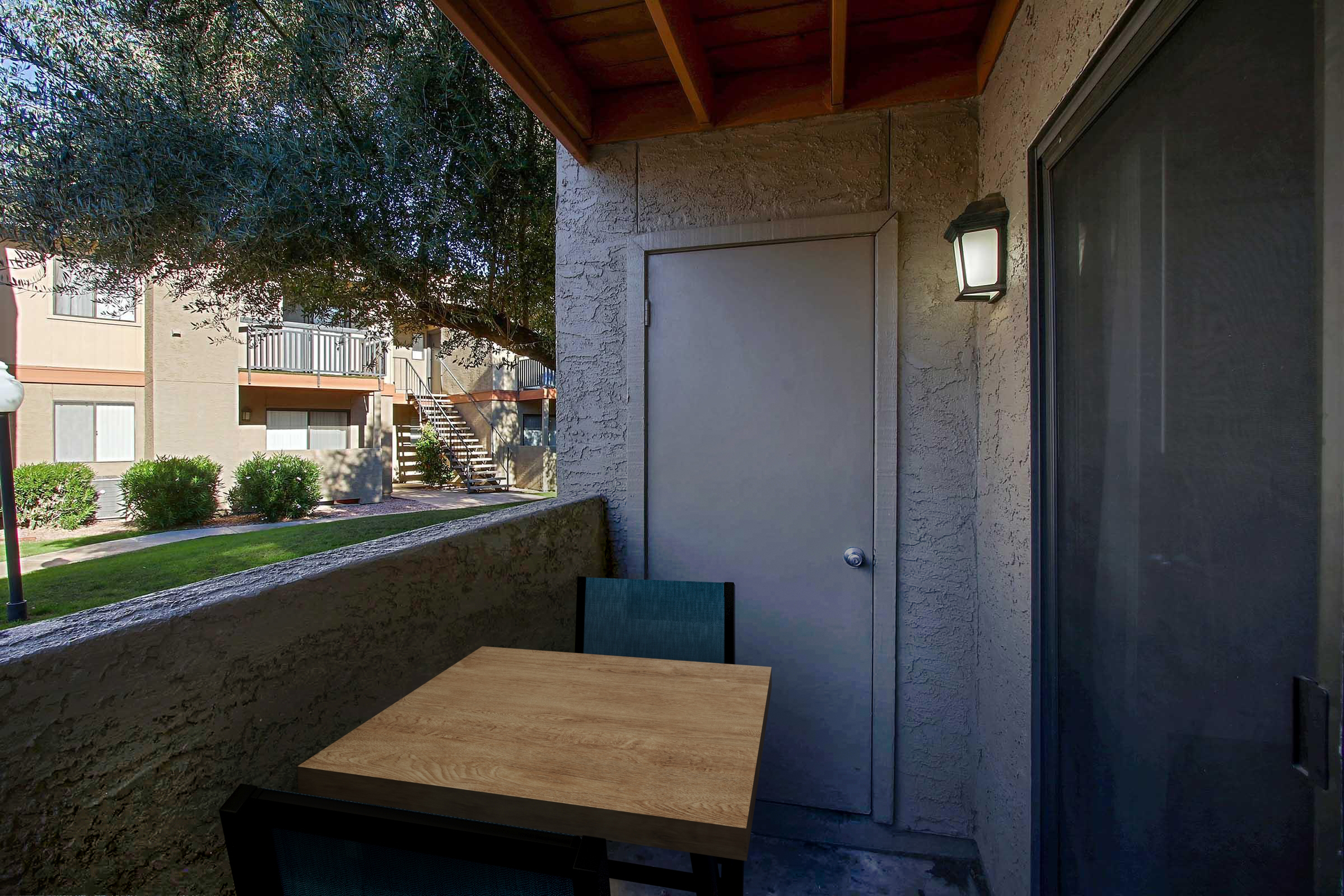A small outdoor patio area featuring a wooden table and two chairs, with a door leading inside. There are green plants and apartment buildings visible in the background, along with a staircase and a streetlight illuminating the entrance.