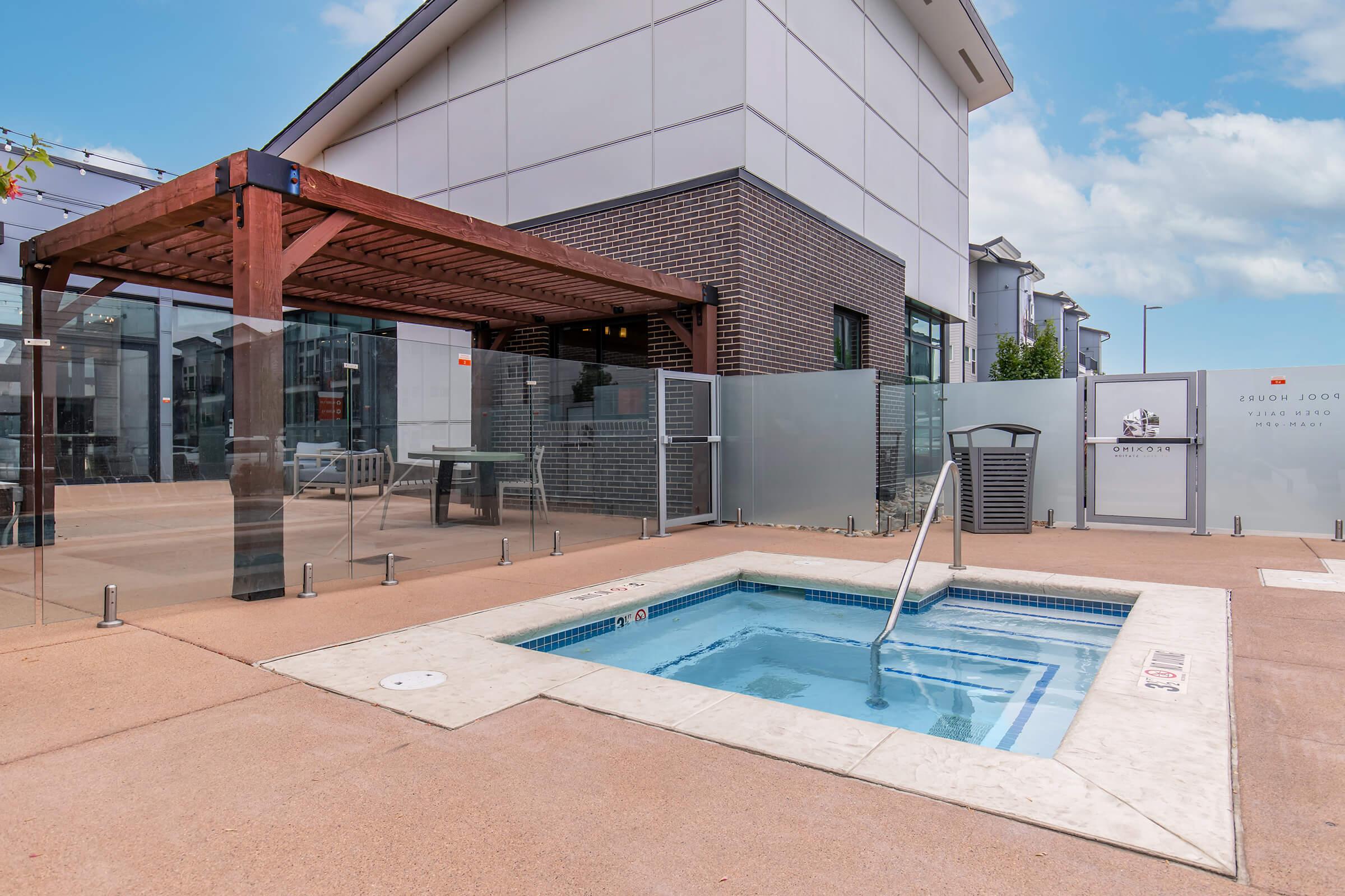 A small outdoor hot tub surrounded by a glass barrier, next to a modern building. A shaded pergola is nearby, along with patio furniture. The area has a clean, contemporary design, with pavers and landscaping in the background under a clear blue sky.