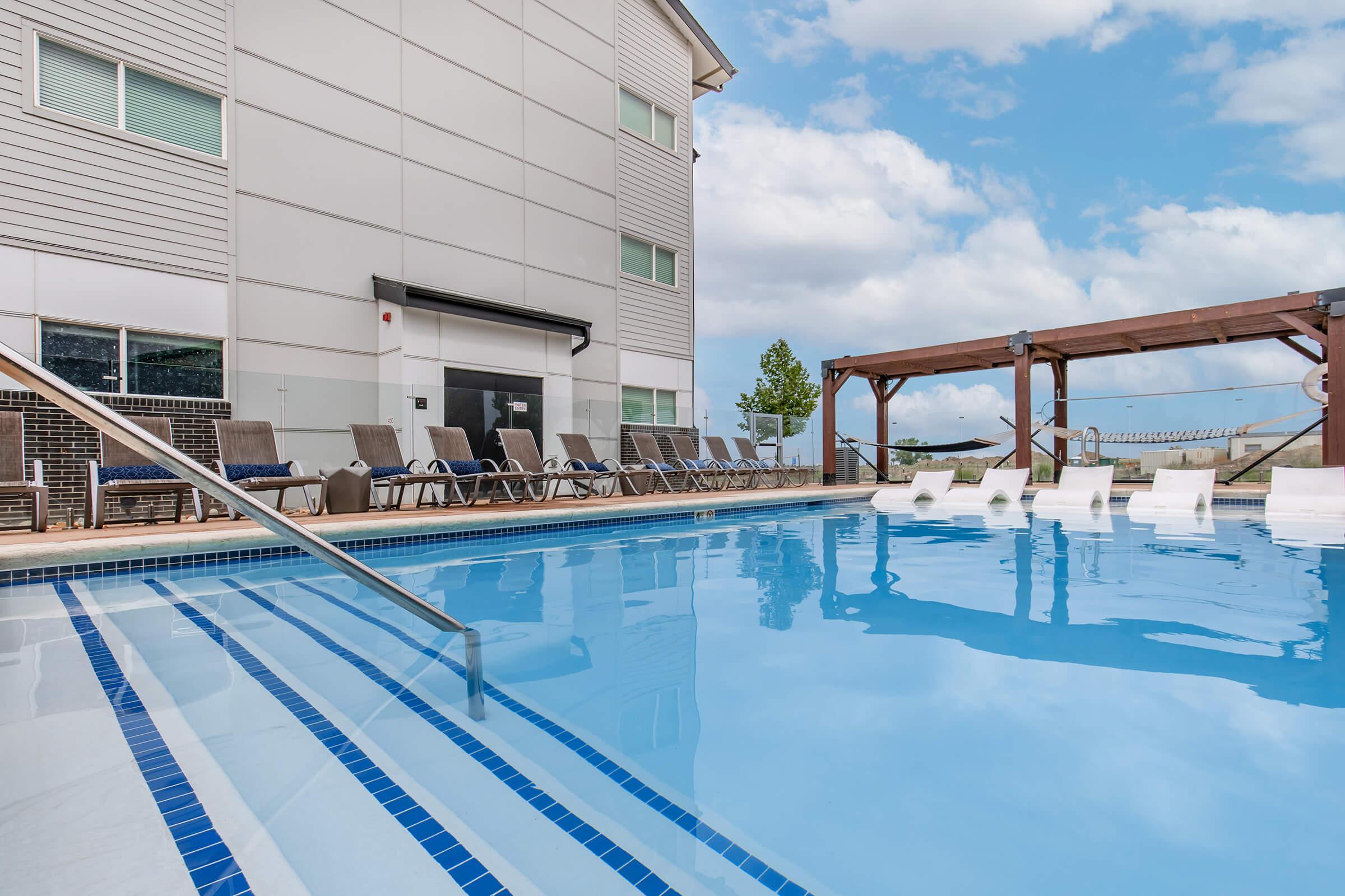 A clear blue swimming pool surrounded by lounge chairs and a wooden pergola. The pool features a staircase entry, and the building nearby has large windows and a modern design. The sky is partly cloudy, creating a serene atmosphere for relaxation and leisure.