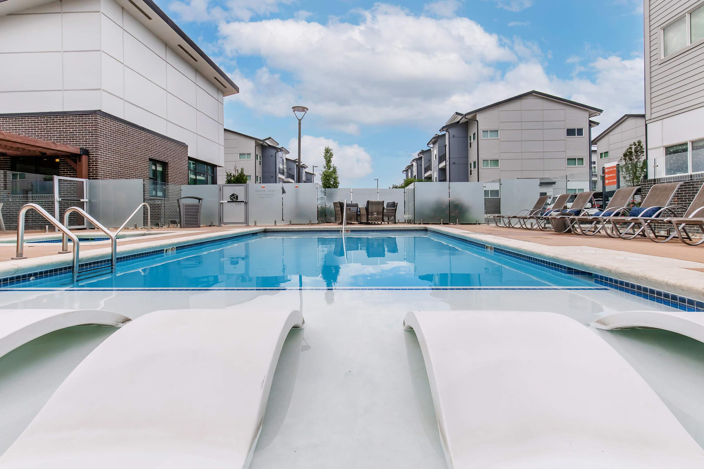 A clear swimming pool with two white lounge chairs in front, surrounded by a deck. In the background, there are modern apartment buildings, a fence, and outdoor seating areas under a cloudy blue sky. The overall scene conveys a relaxing outdoor environment.