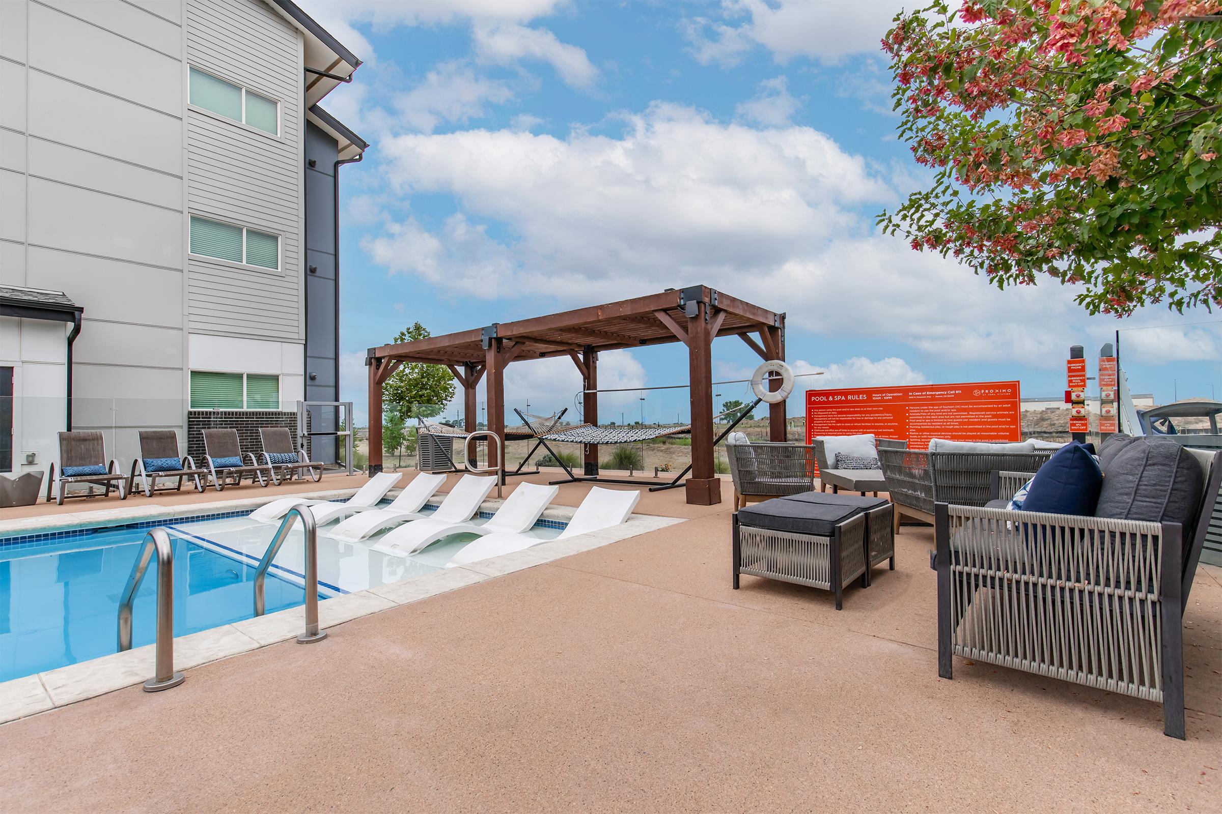 A pool area featuring a clear blue swimming pool with lounge chairs lined up beside it. There’s a wooden pergola with a seating area and shaded spots, along with a vibrant orange wall displaying information or signage. Lush trees and a partly cloudy sky add to the serene ambiance.