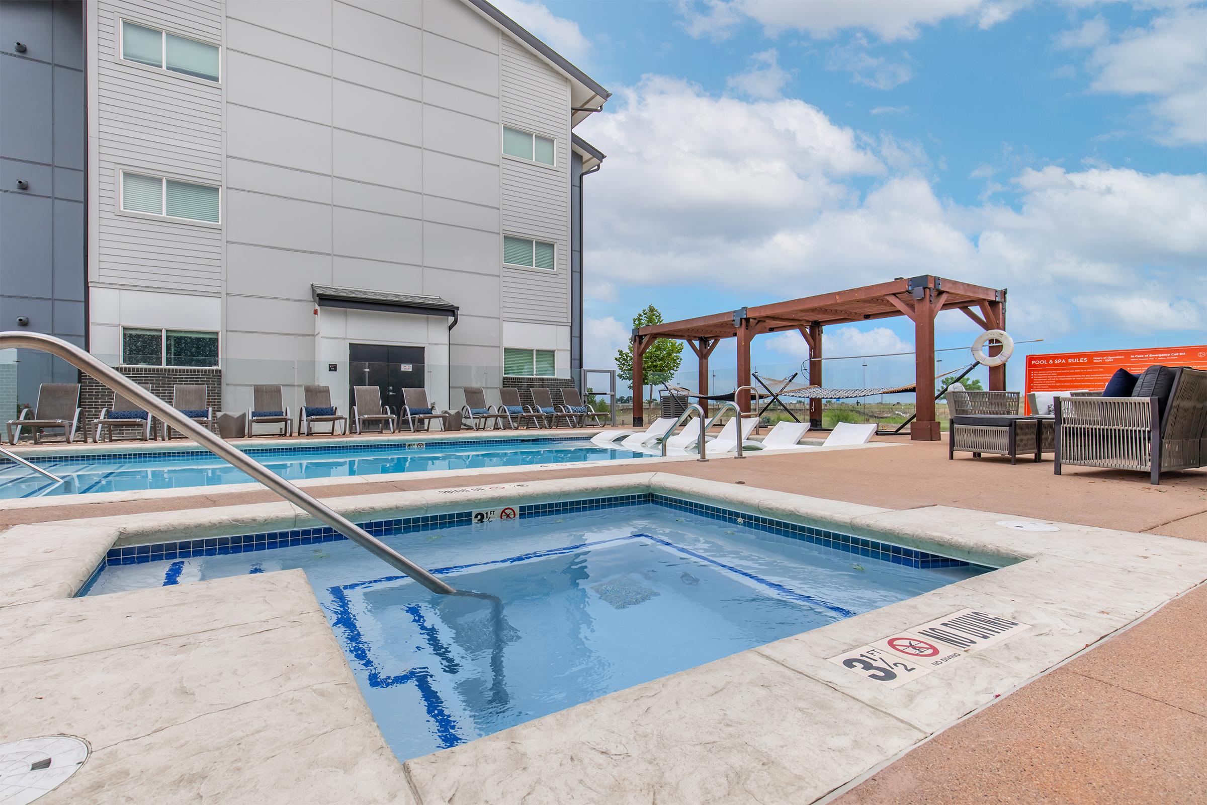 A spacious outdoor pool area featuring a large swimming pool with lounge chairs arranged around it. There is a hot tub nearby, along with a shaded seating area under a wooden pergola. The background includes a modern building and a clear blue sky.