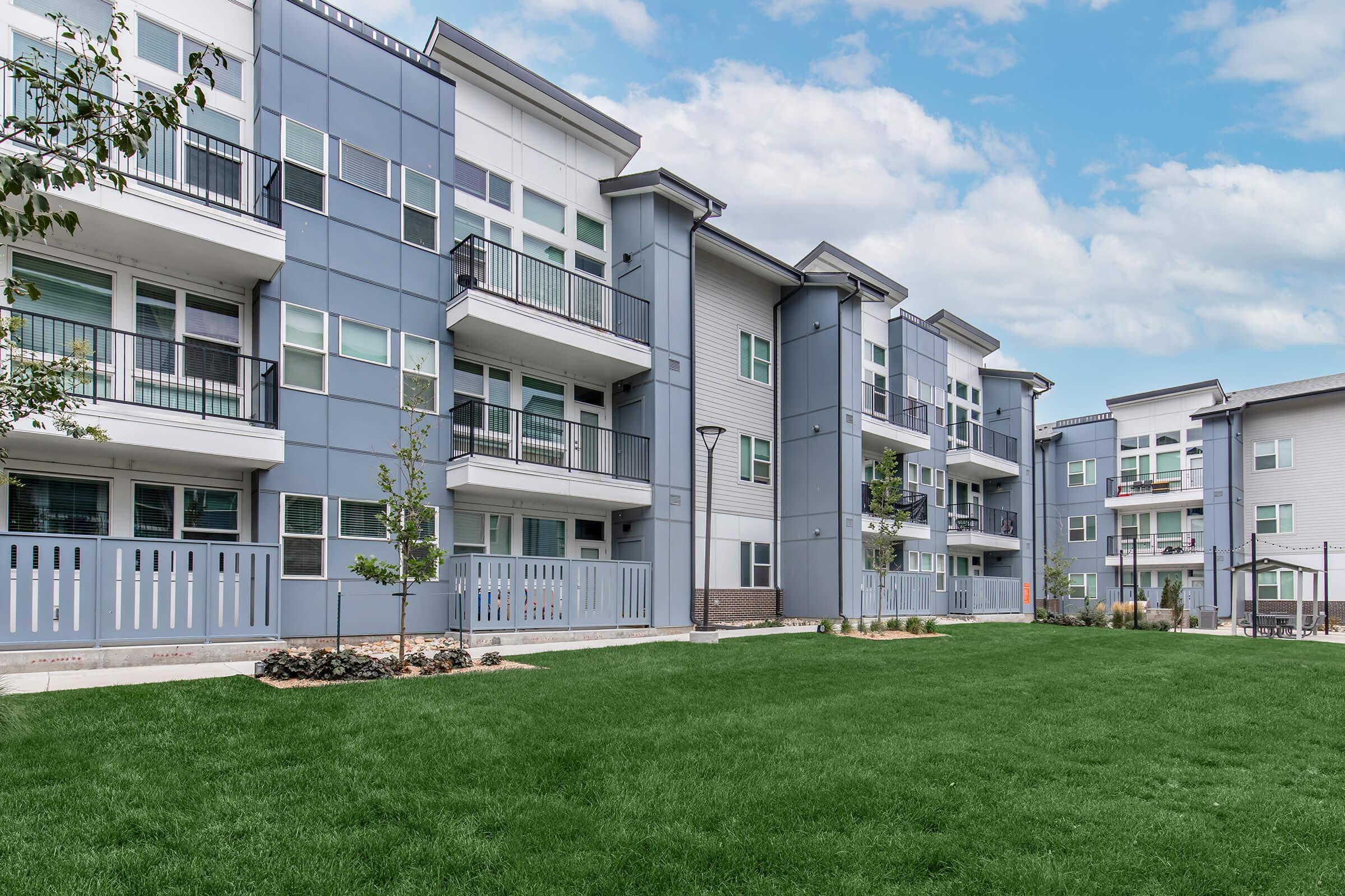 A modern apartment complex featuring multiple stories with a mix of blue and grey siding. Balconies are visible on several units, and the landscaped area includes well-maintained green grass and small trees. The sky is partly cloudy, adding to the bright, inviting atmosphere of the outdoor space.