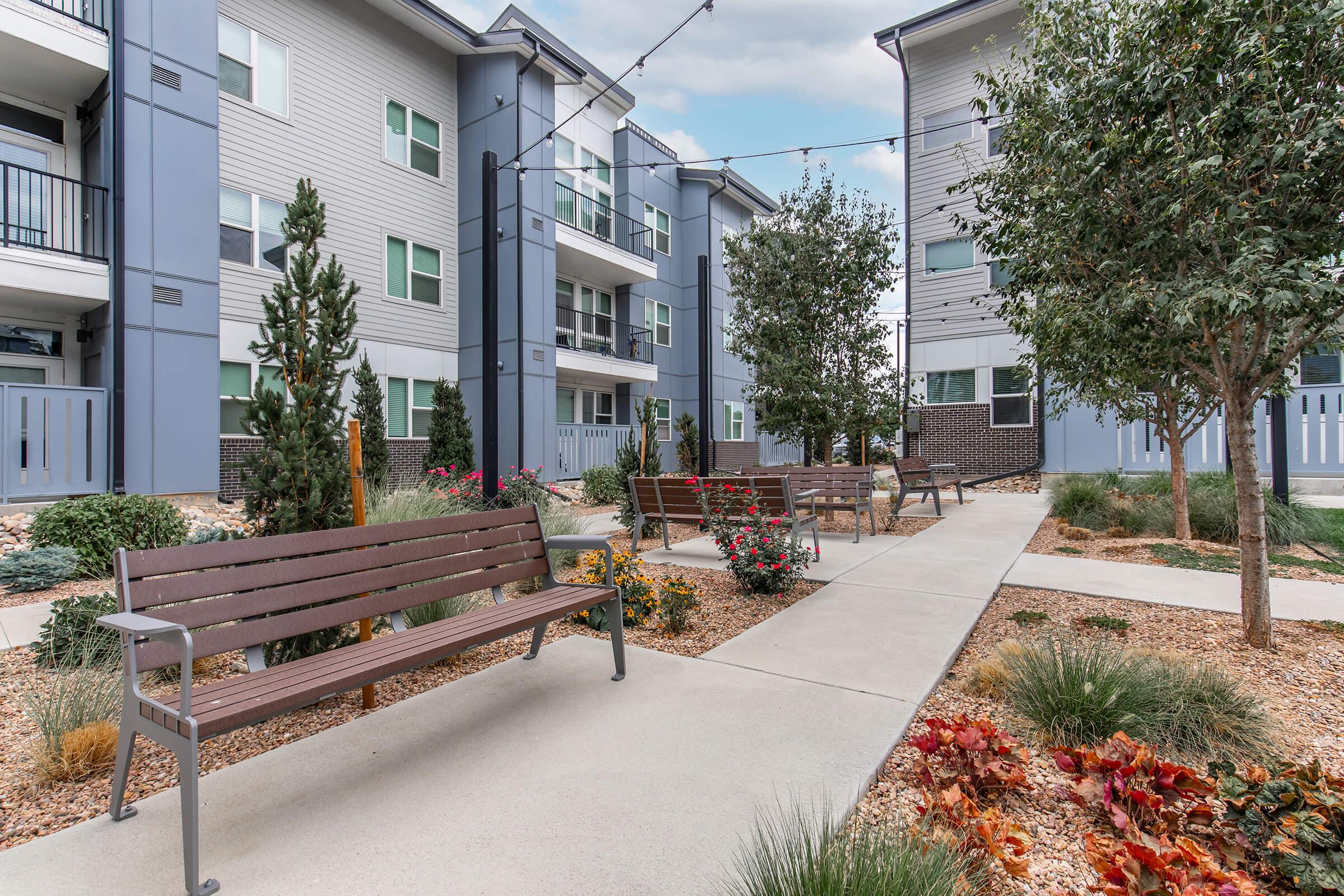 A modern residential courtyard featuring landscaped gardens, benches, and walking paths. The area is surrounded by multi-story apartment buildings with exterior lighting, shrubs, and decorative plants, creating an inviting and peaceful outdoor space.