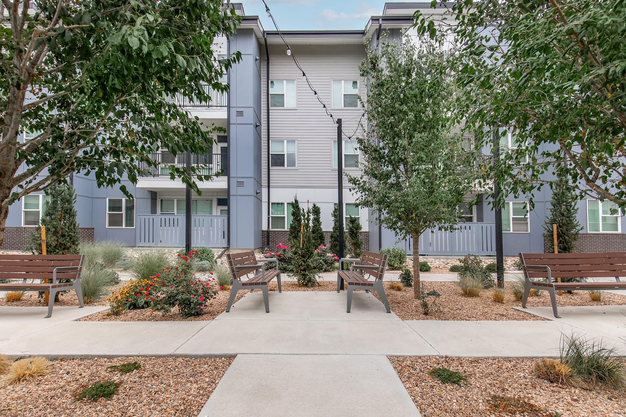 A landscaped courtyard featuring flower beds and benches, surrounded by modern apartment buildings with gray siding. The area is lined with young trees and decorative shrubs, creating a welcoming outdoor space for residents. Pathways run through the garden, connecting different sections of the courtyard.