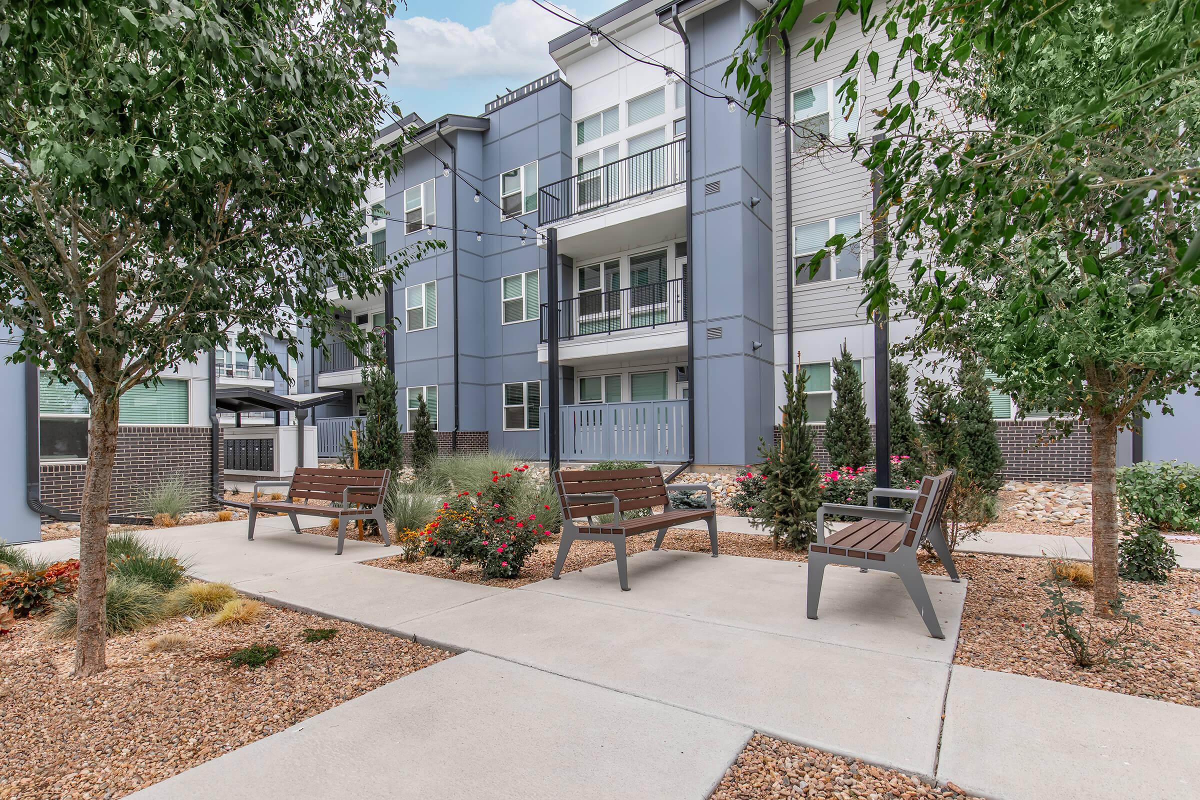 A modern multifamily apartment building with a gray exterior, featuring landscaped pathways lined with benches, shrubs, and flowering plants. The scene is bright and inviting, with a focus on outdoor seating areas for residents.