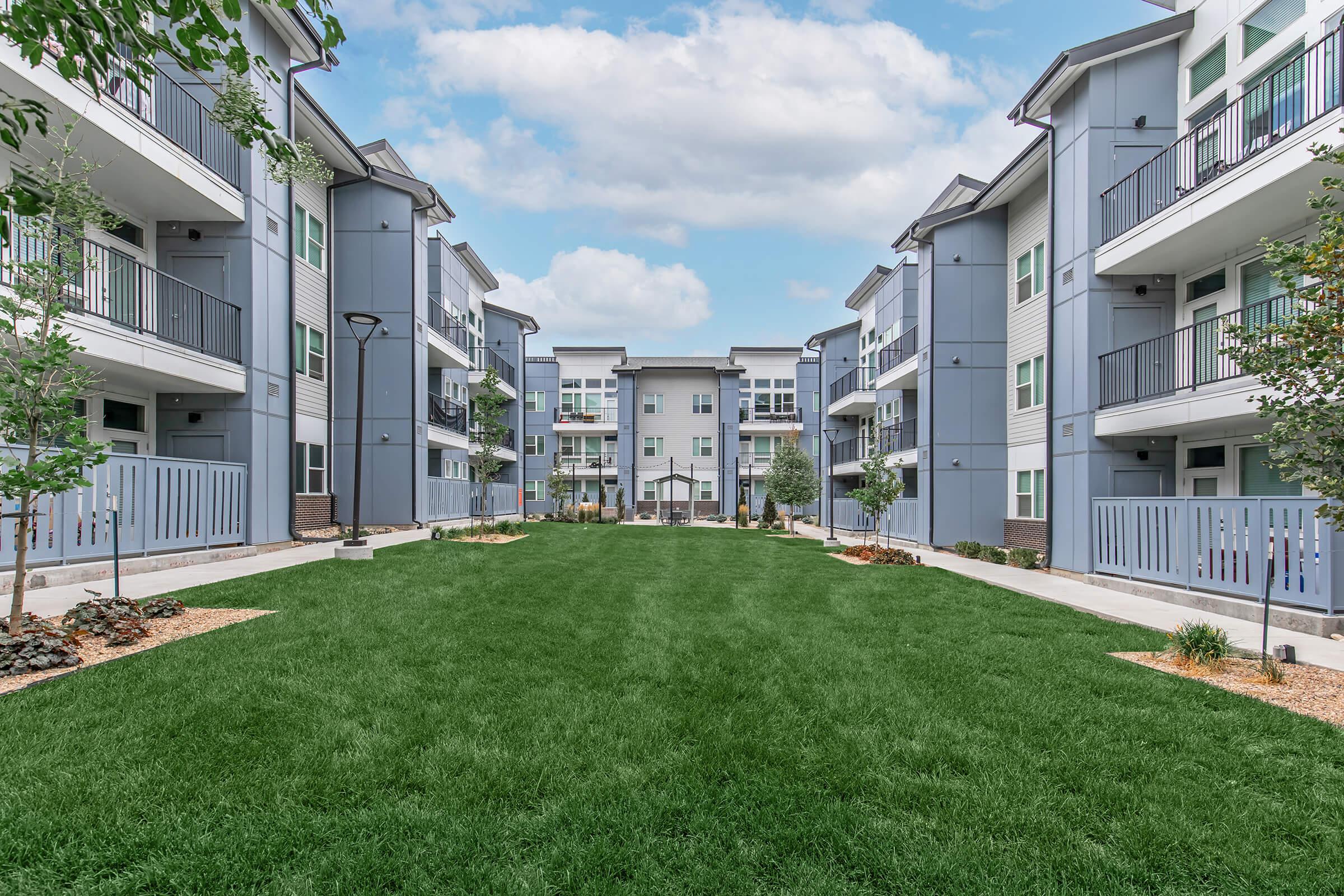 A view of a modern apartment complex with two rows of buildings on either side. The center features a green lawn bordered by decorative landscaping. There are balconies visible and well-maintained walkways. The sky is partly cloudy, creating a bright and inviting atmosphere.