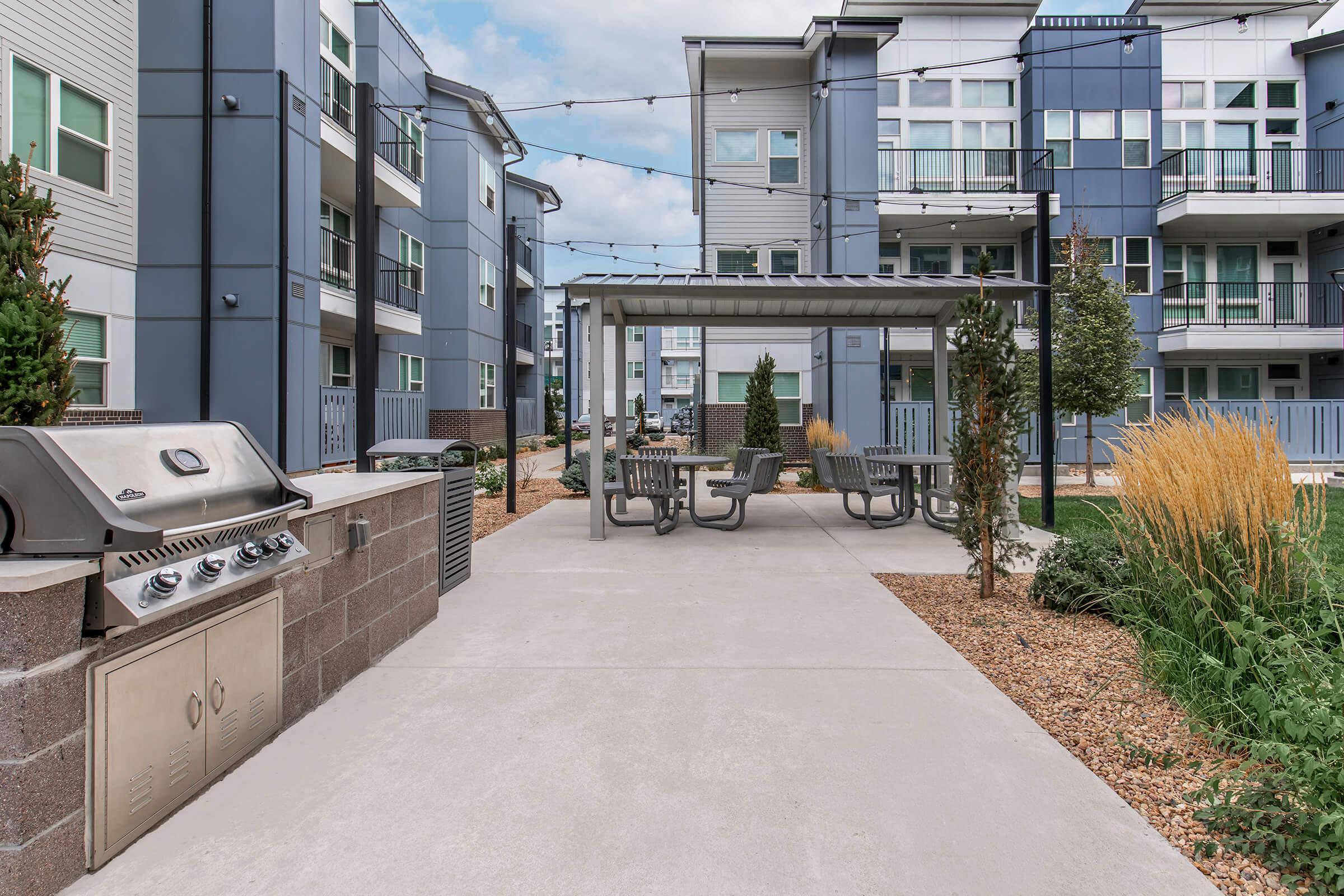 Outdoor recreational area featuring a concrete pathway leading to a covered patio with tables and chairs. A grill is set up nearby, surrounded by landscaped gardens and modern apartment buildings in the background. String lights are hung above the seating area, creating a cozy atmosphere.